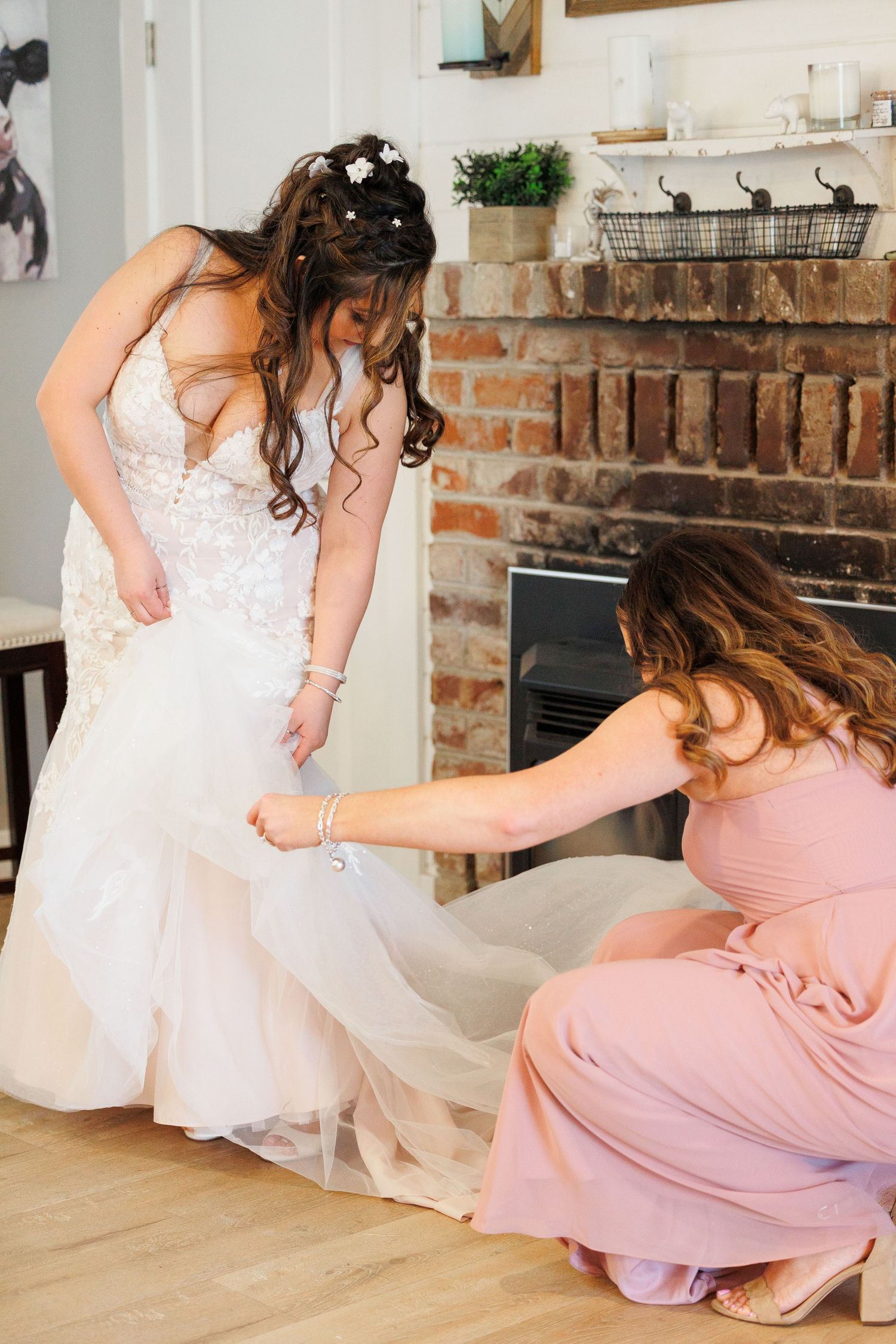 Person in pink dress helps adjust white wedding gown train on wooden floor near exposed brick wall.