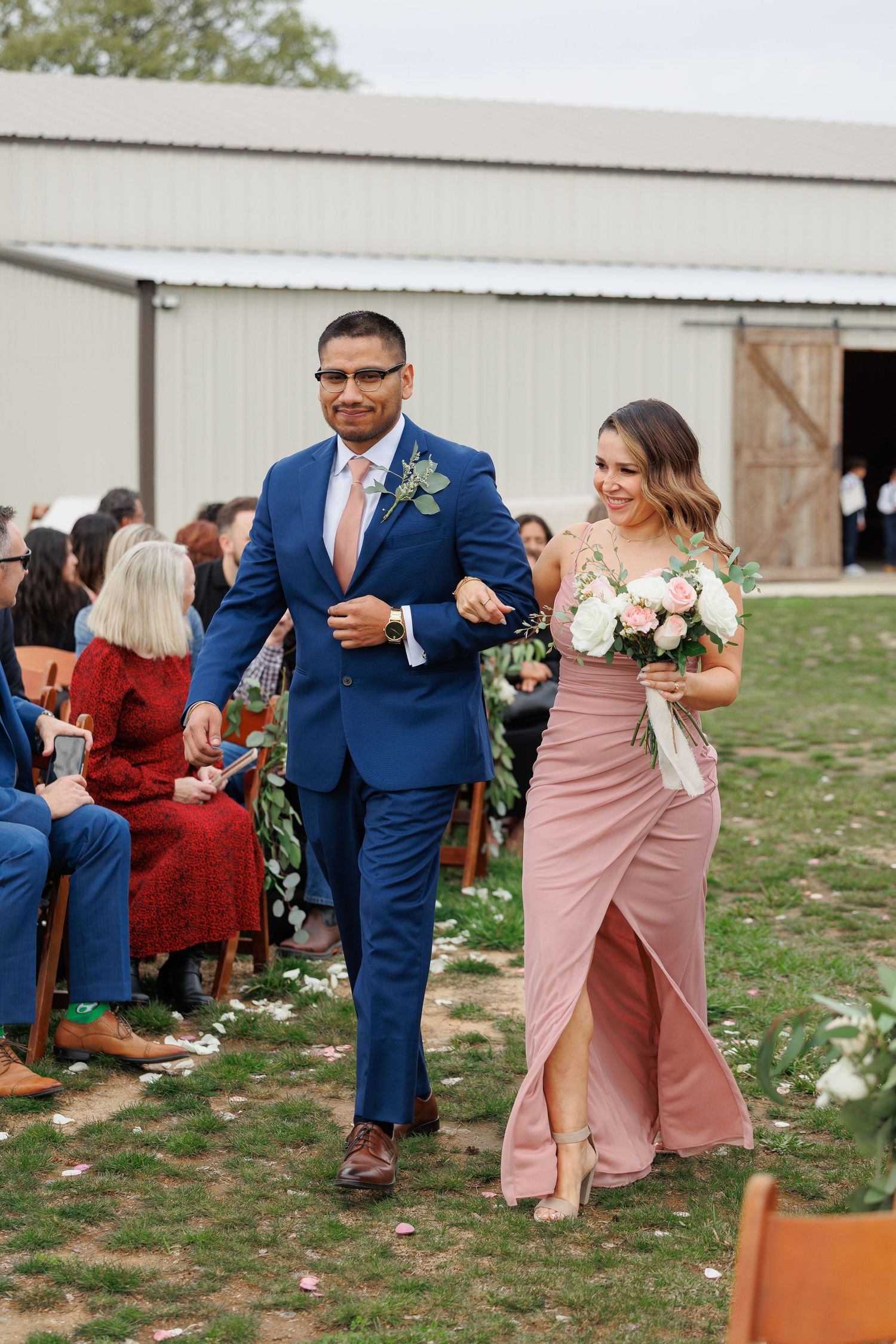 Wedding party members walk arm in arm down outdoor aisle in navy suit and dusty rose dress at rustic barn venue.