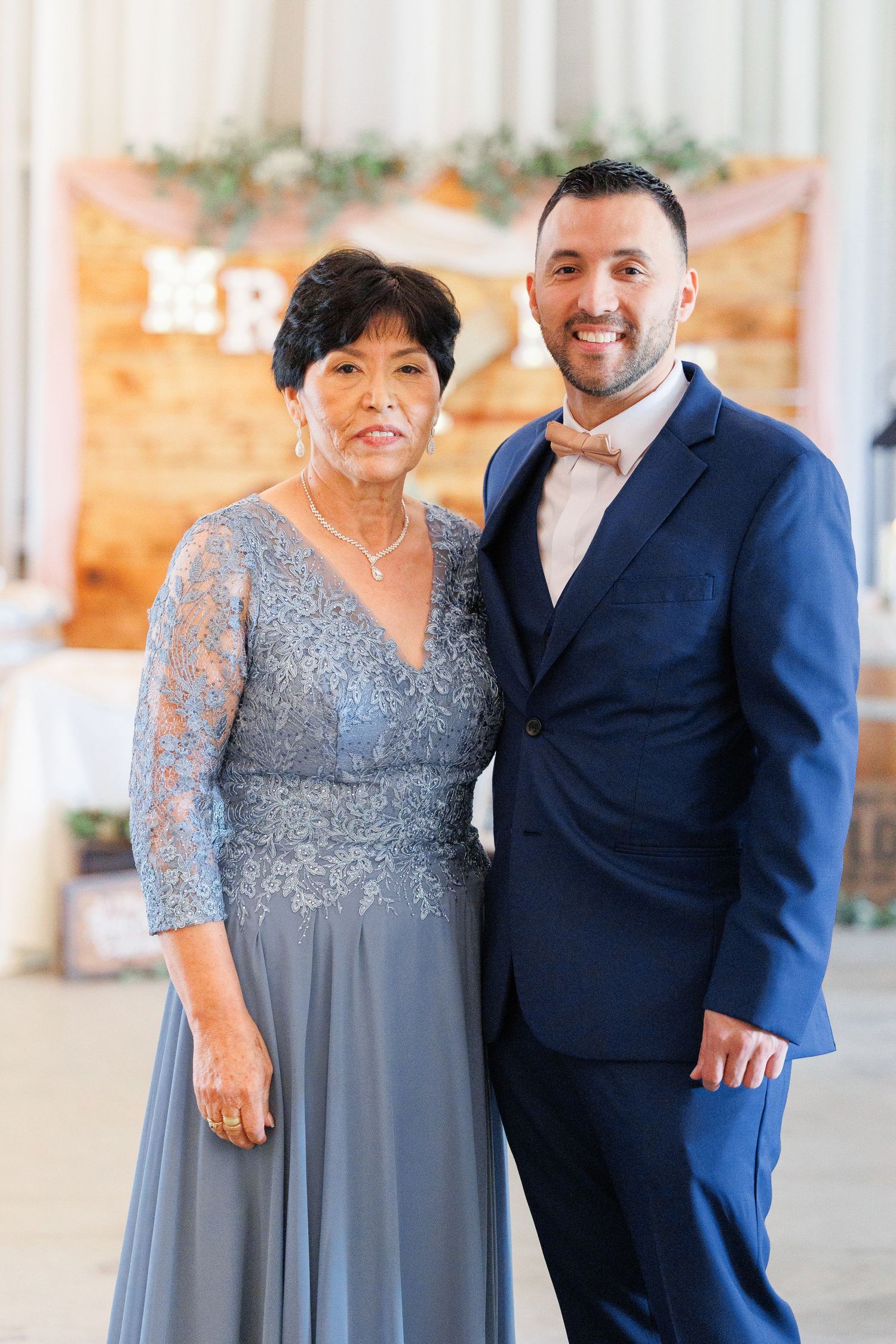 Two people in formal attire pose together at an event, with one wearing a blue suit and the other in a gray lace gown.