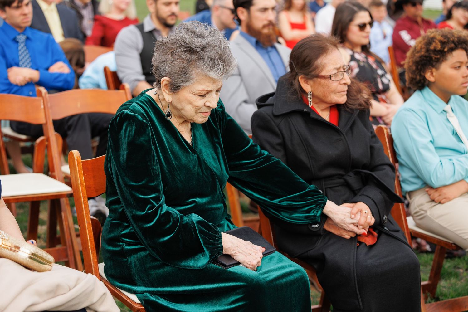 Attendees sit in wooden chairs at an outdoor event with someone wearing an elegant emerald green velvet dress.