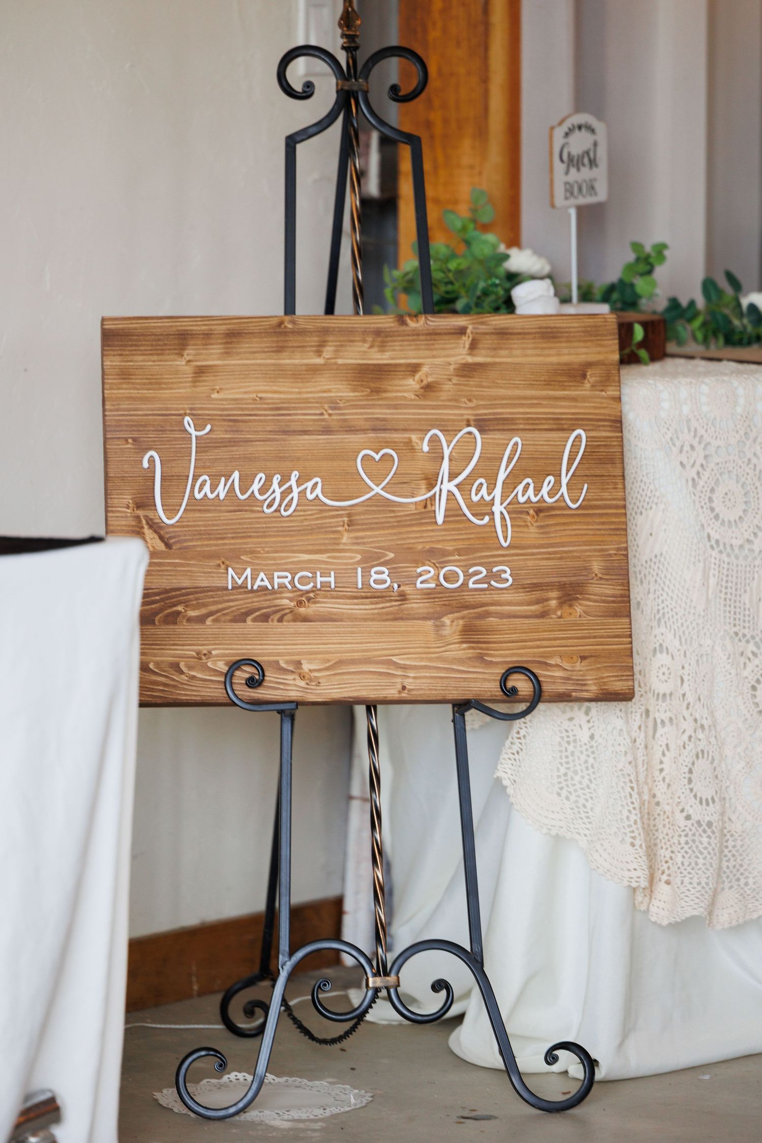 Rustic wooden wedding signs with white calligraphy text displayed on decorative iron stands against white lace backdrop.