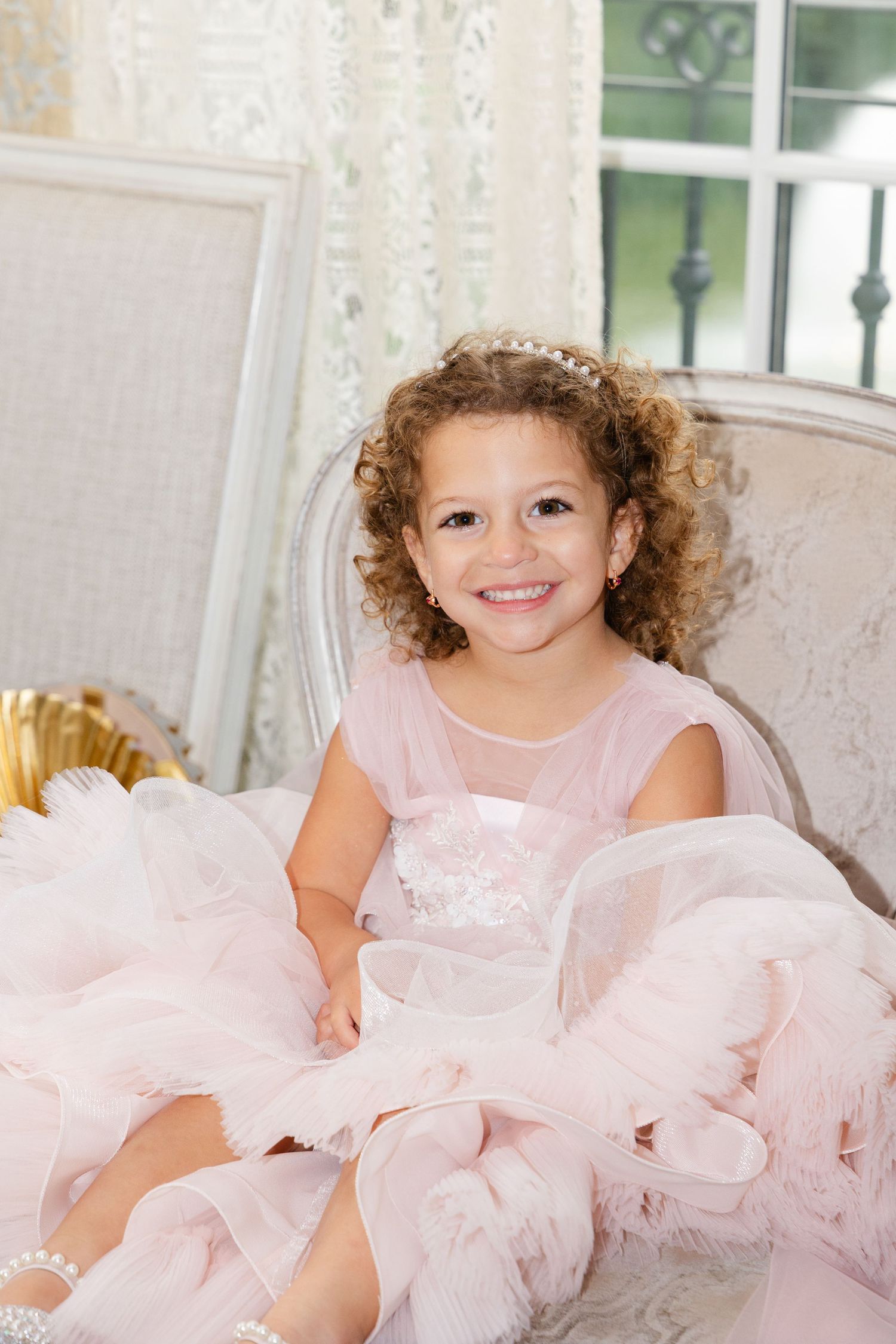 A young child in a pink tulle dress poses on a white couch with a bright smile.