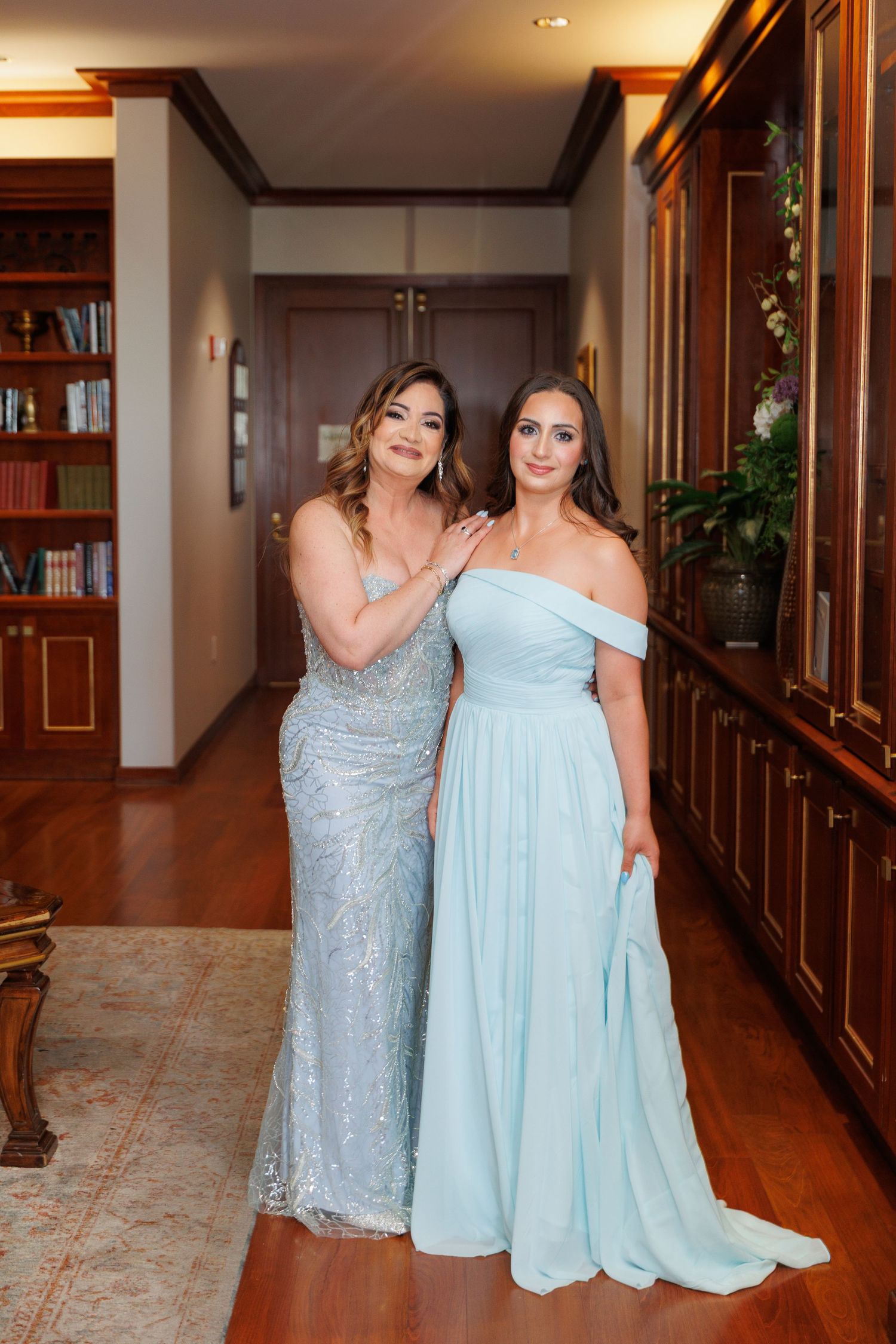 Two women in elegant evening gowns, one in sparkly silver and one in light blue, pose together in a hallway.
