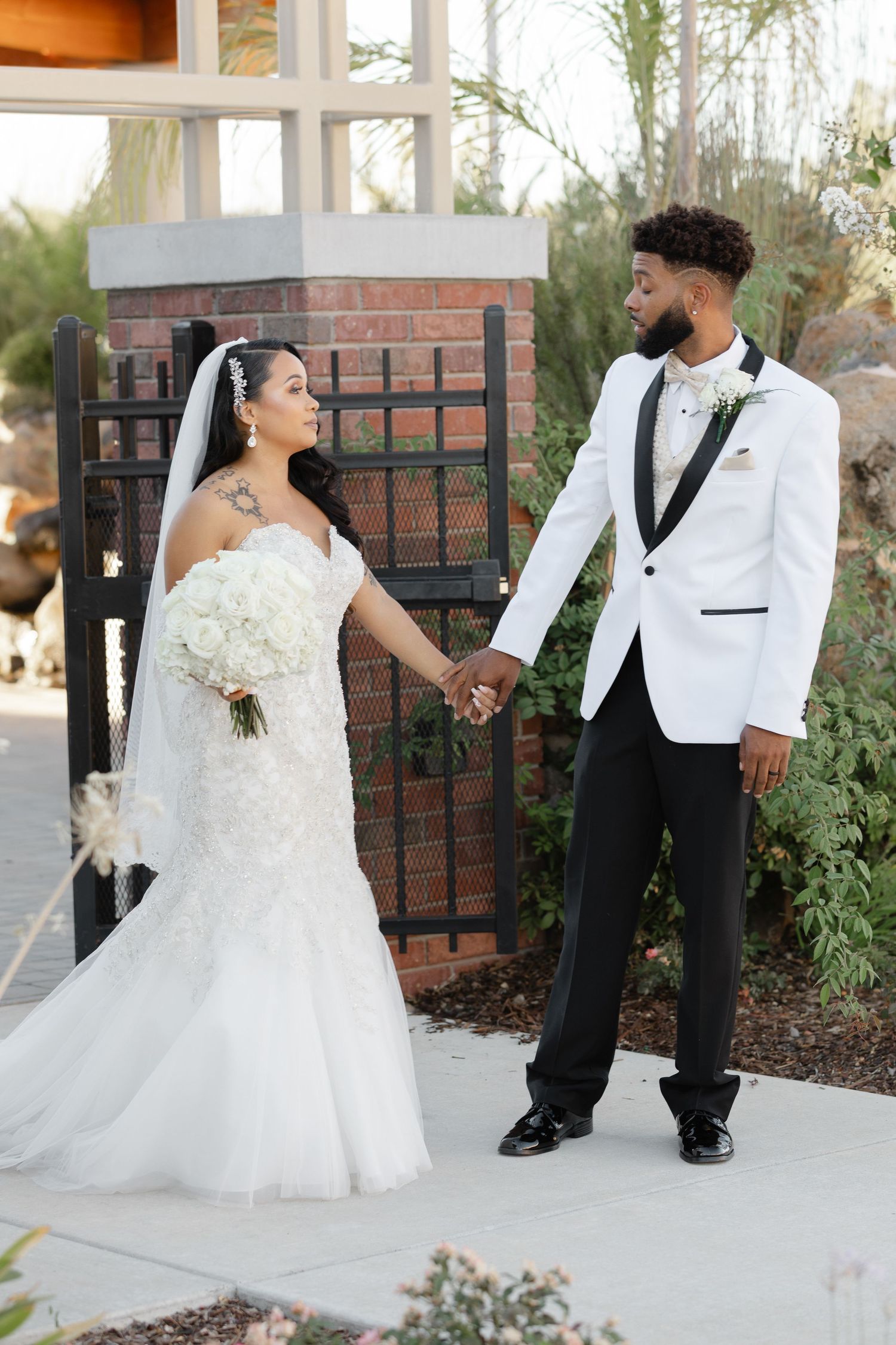 A romantic wedding couple poses together in front of a brick wall, with the bride wearing a flowing white gown.