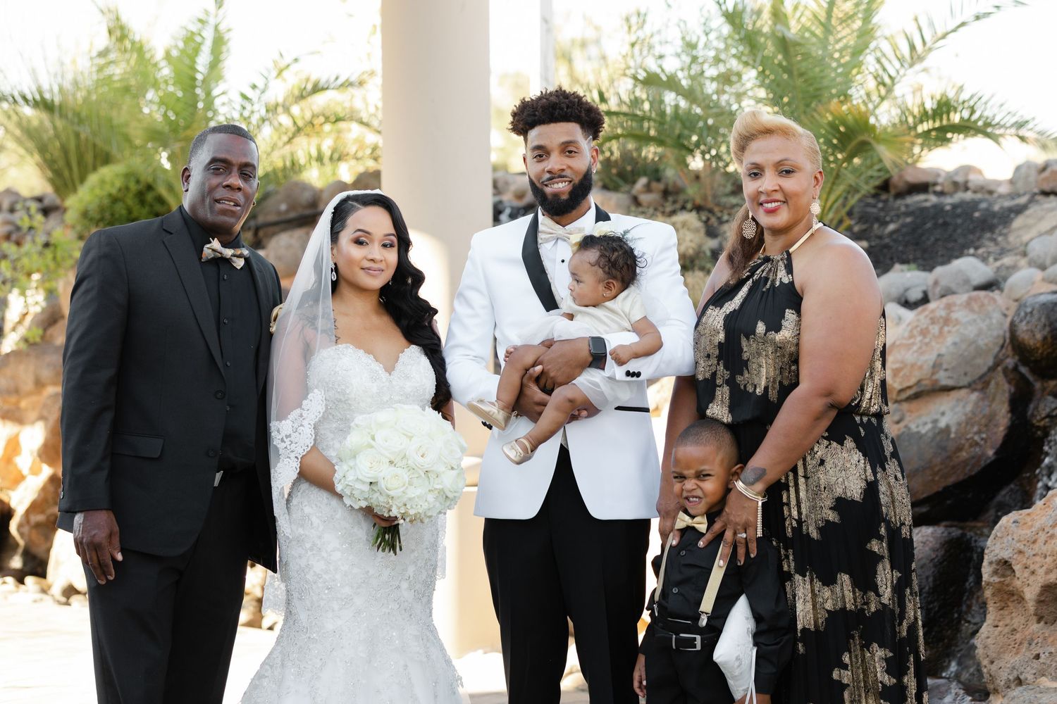 A wedding party poses together outdoors with palm trees and rocky backdrop on a sunny day.
