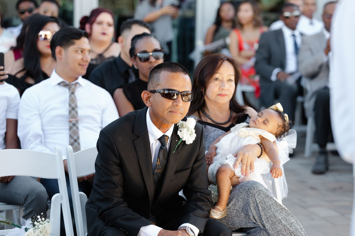 Wedding guests in formal attire seated in white chairs during an outdoor ceremony at sunset.
