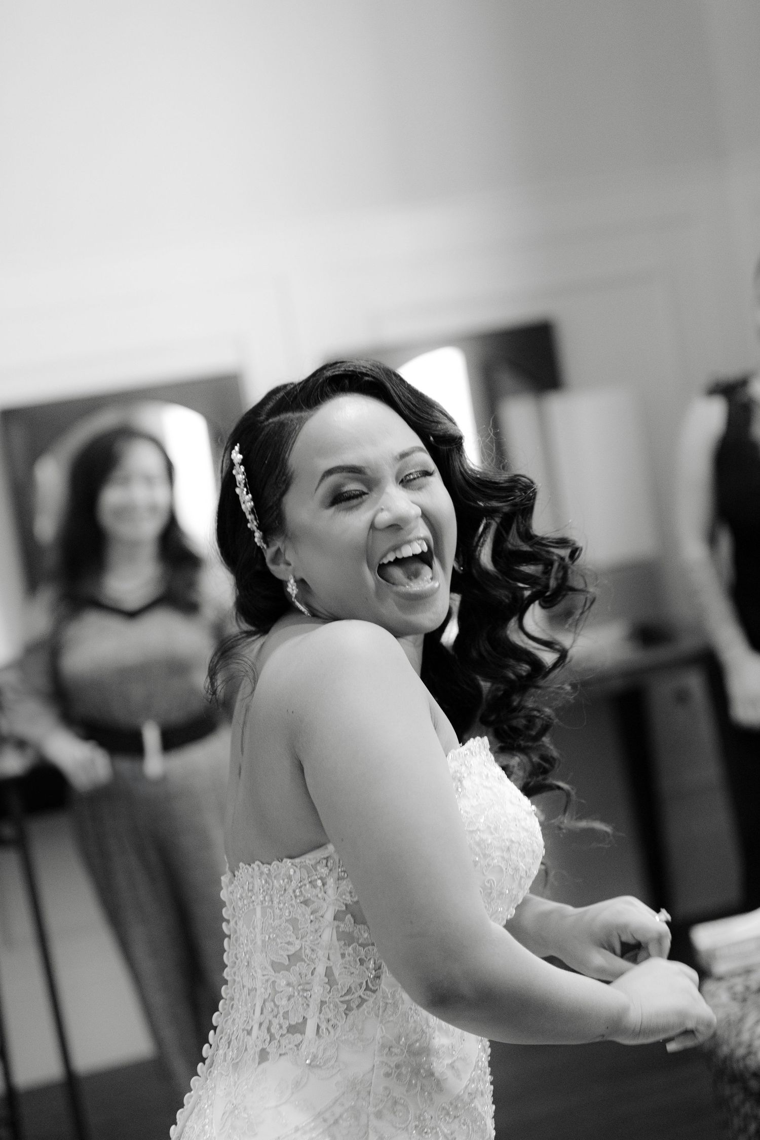 A joyful bride in a white lace wedding dress laughs with curled dark hair and natural makeup in black and white photo.