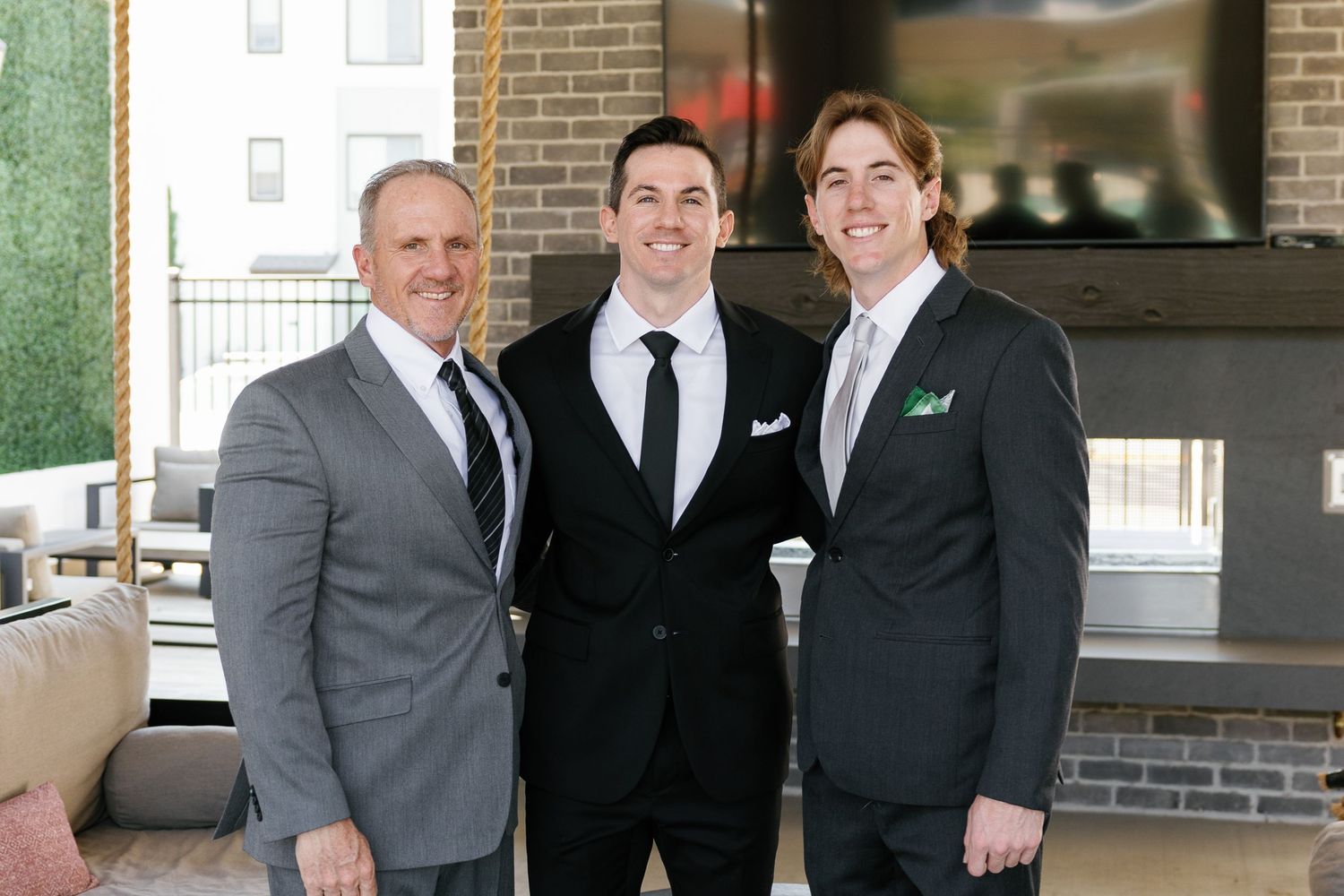 Three men in formal suits stand together smiling at an indoor venue with brick walls and natural lighting.