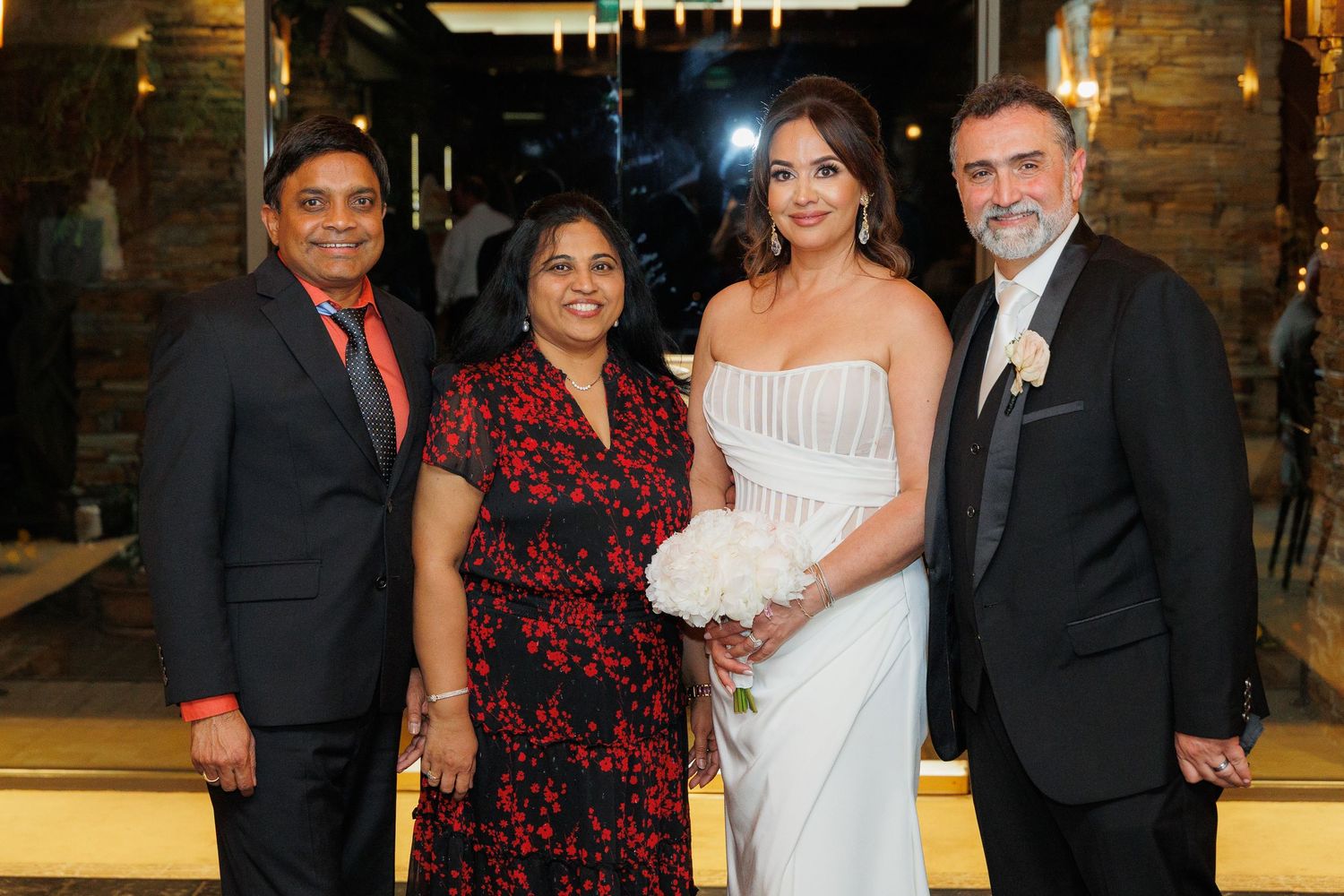 Four people stand together at a formal event, with two wearing dark attire and one in an elegant white gown.