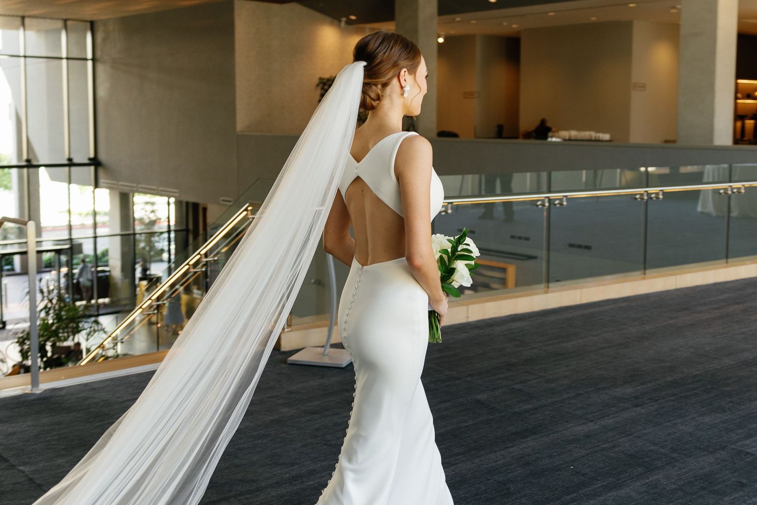 Bride in a flowing white wedding dress and veil walks through a modern interior hallway.