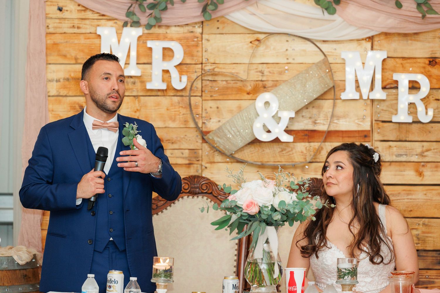 Wedding couple and guests share joyful moments at a rustic wooden barn reception venue decorated with greenery.
