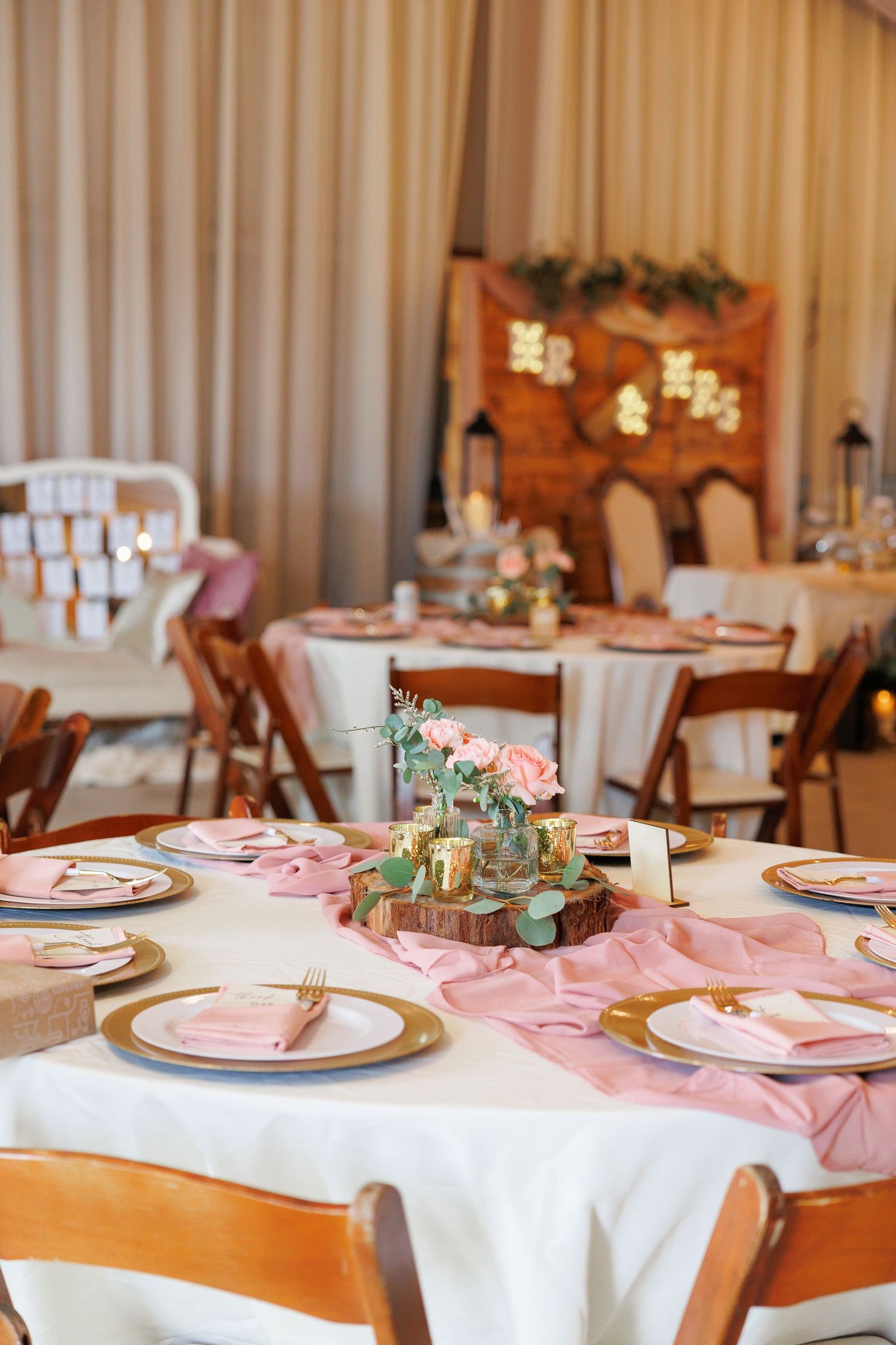 Elegant wedding reception table setting with pink napkins, gold charger plates, and rustic wooden chairs.