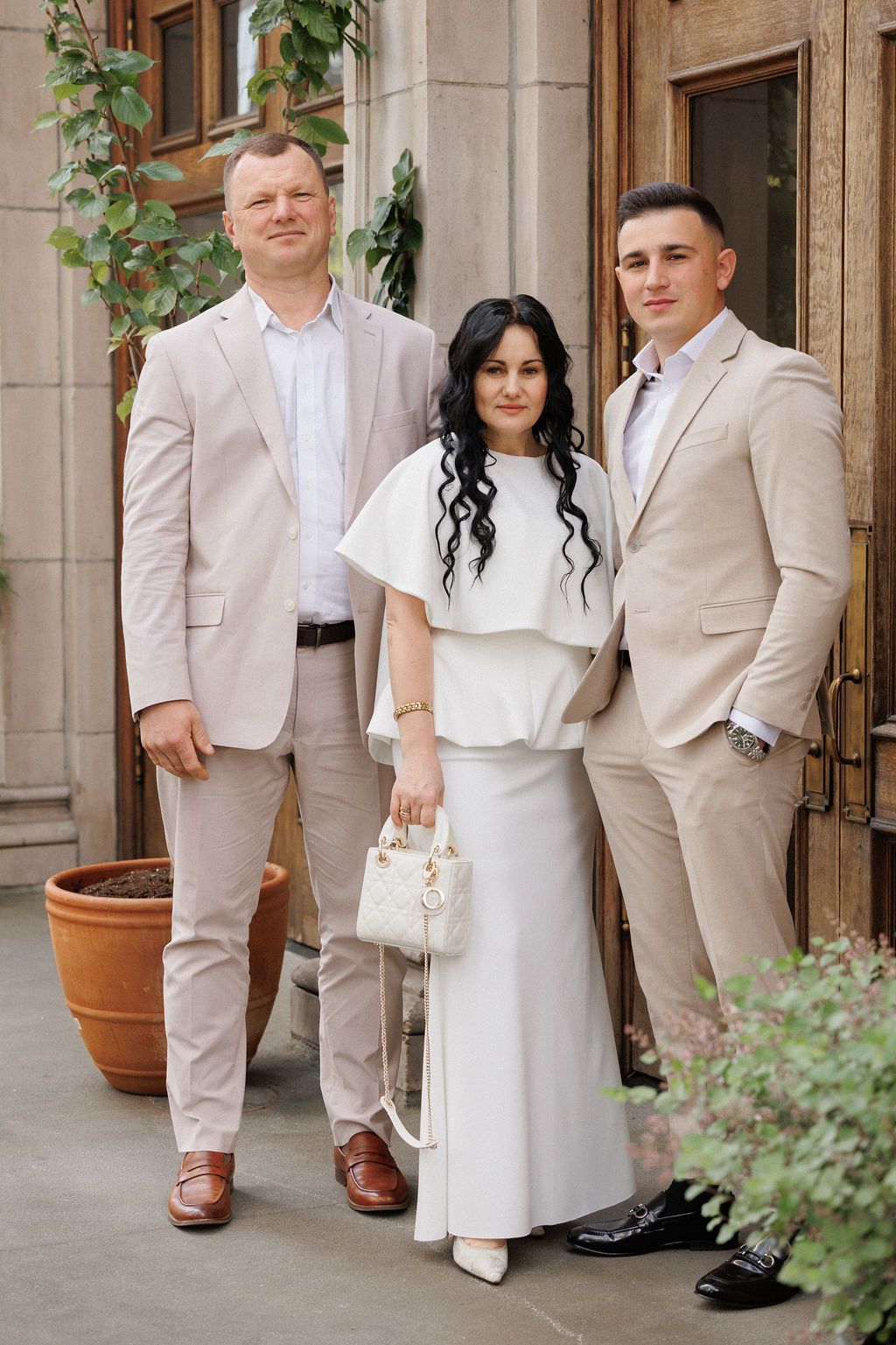 A wedding party poses outdoors in coordinated beige and white attire against a stone building with greenery.
