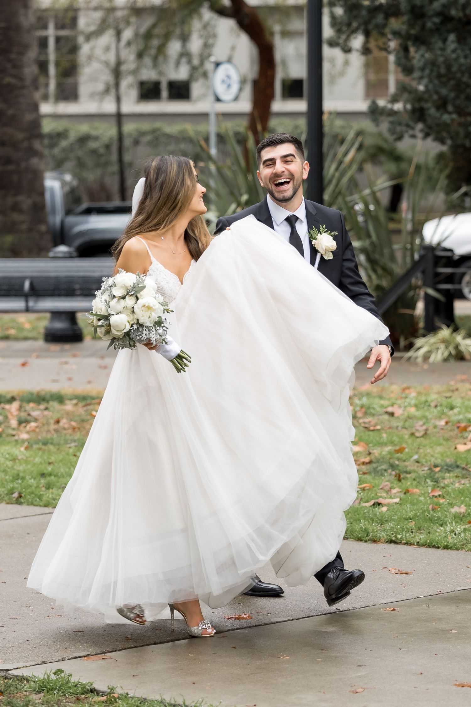 A joyful bride and groom share a playful moment running and spinning together on a leaf-covered sidewalk.