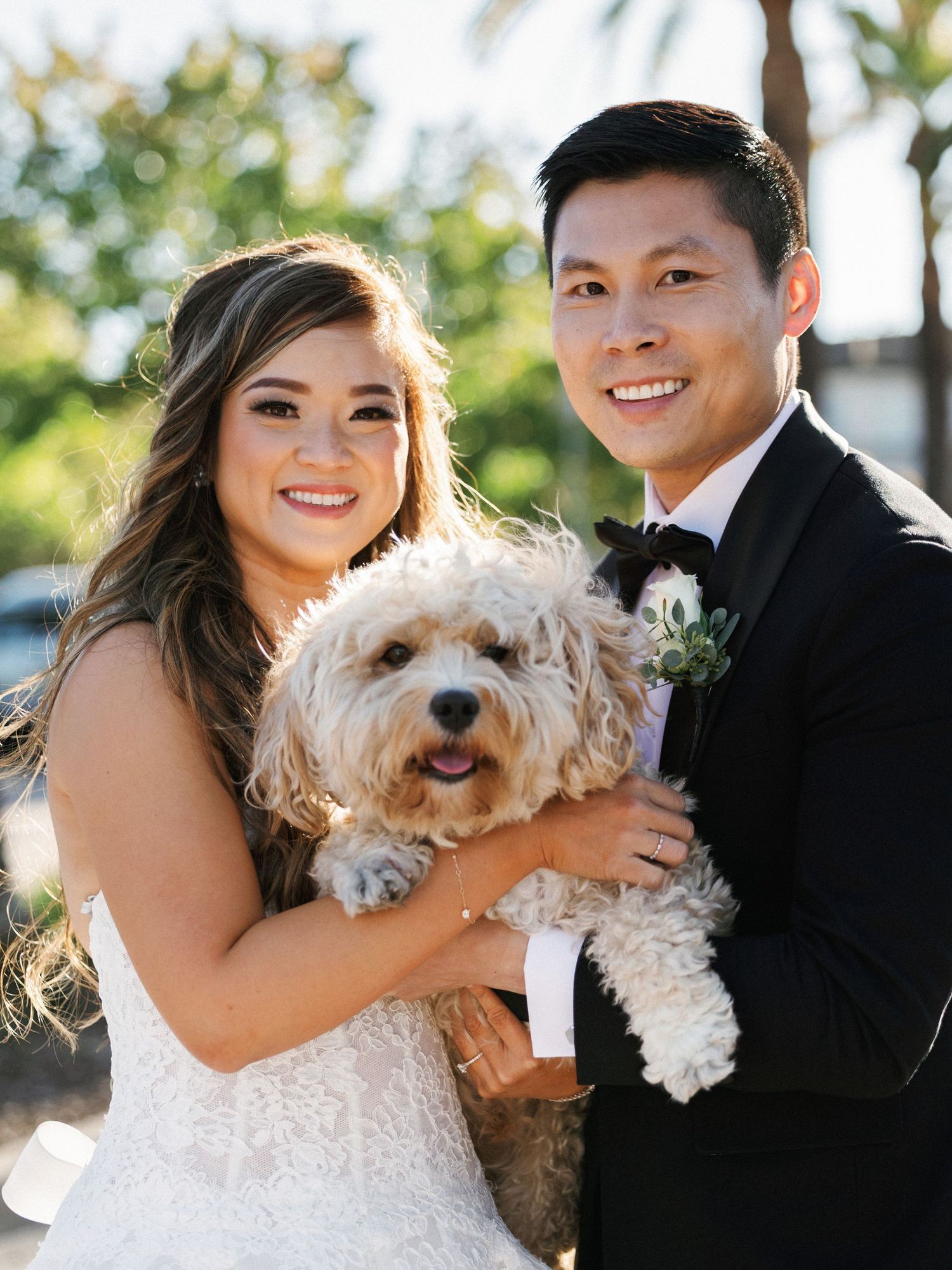 A fluffy white dog joins the couple for outdoor wedding portraits under natural sunlight.