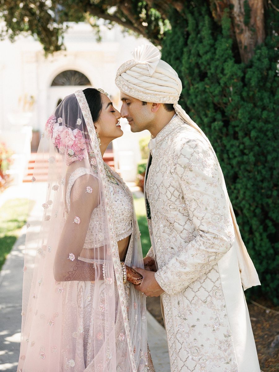 A romantic wedding couple in traditional South Asian attire embraces outdoors against a green garden backdrop.