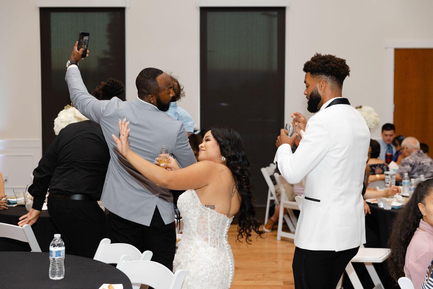 Wedding party members dance and celebrate together during a reception in a bright indoor venue.
