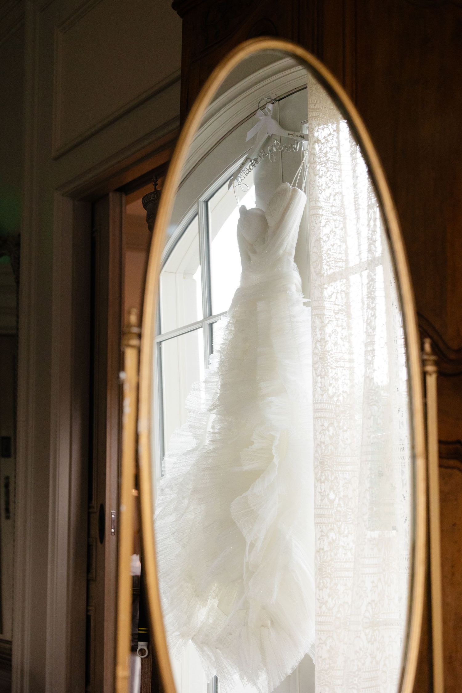 A white wedding dress hangs in an arched doorway with natural light streaming through, creating a romantic silhouette.