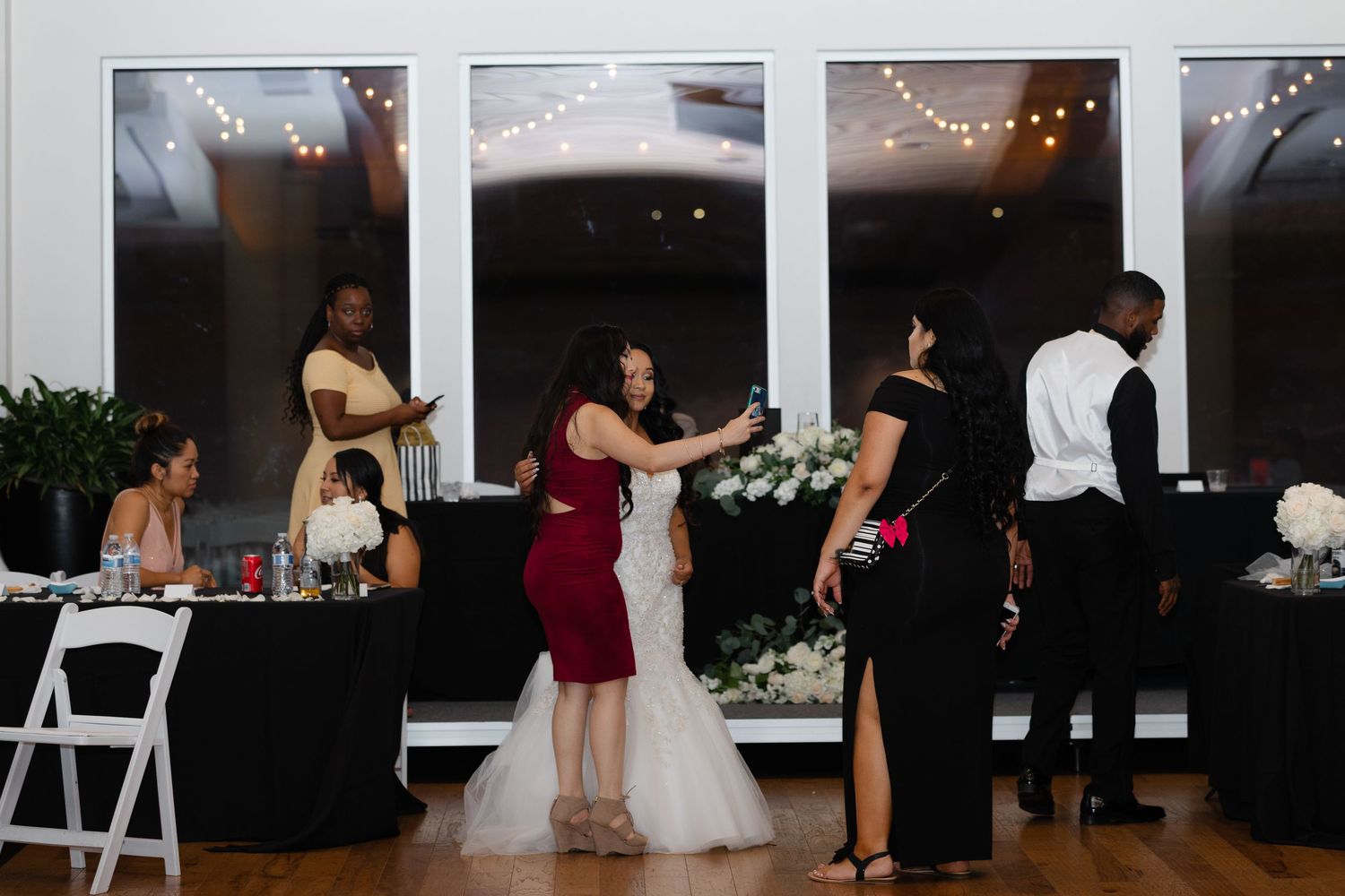 Wedding reception guests gather at a dimly lit venue with string lights overhead as people dance and mingle near the buffet area.