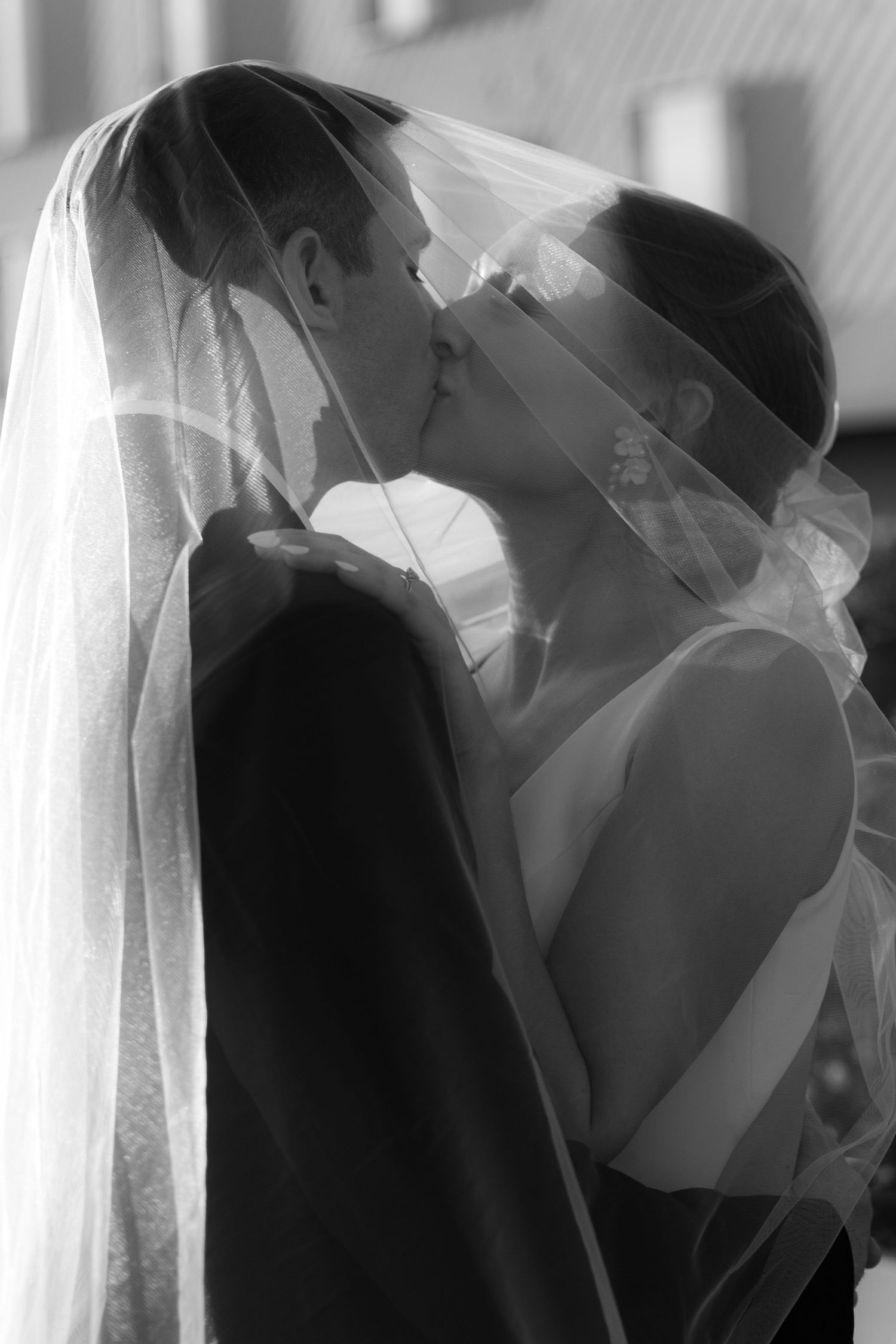 A romantic black and white wedding photo of a couple sharing their first kiss under a flowing veil.