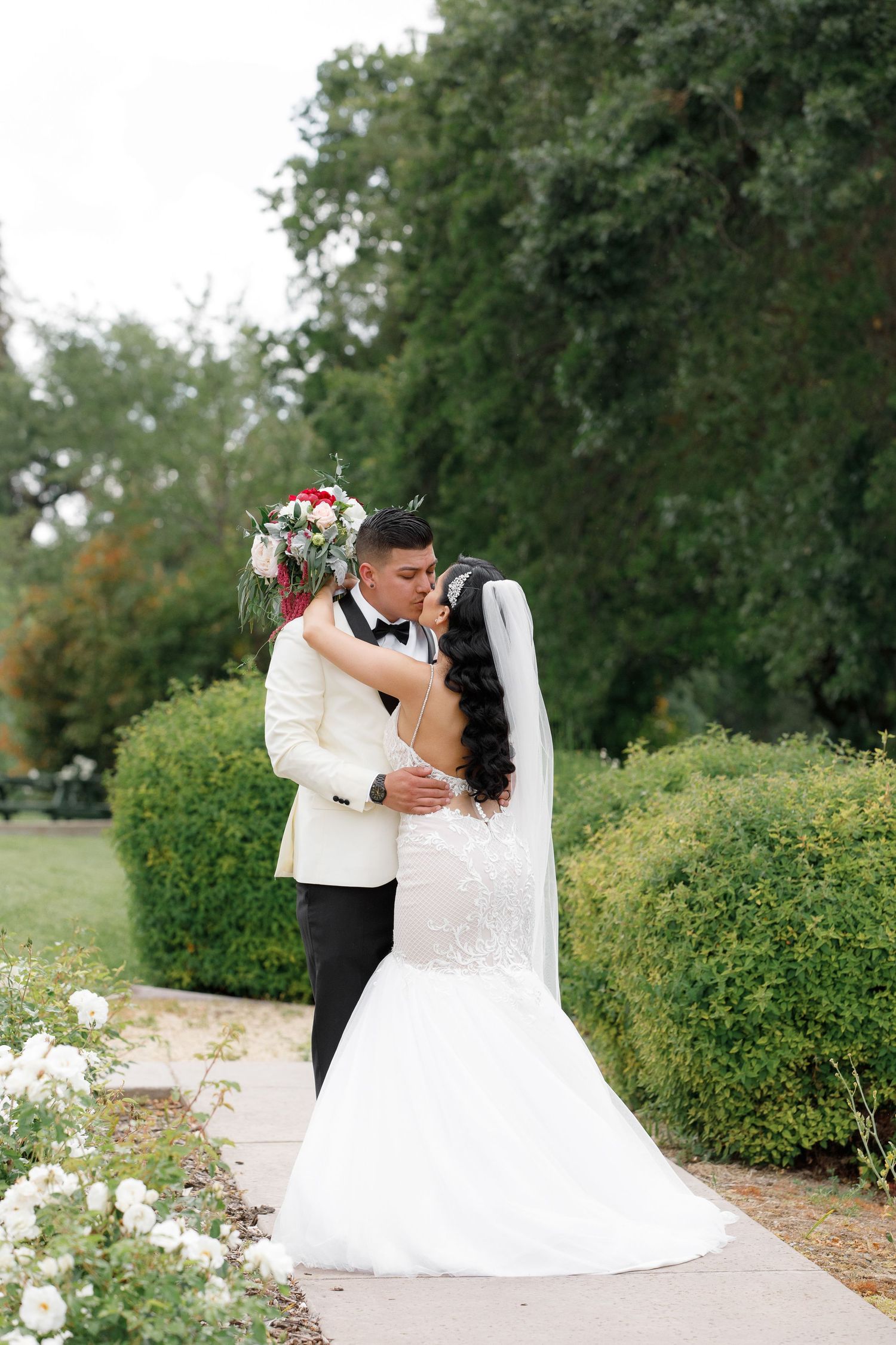 A romantic outdoor wedding scene with a couple embracing in a garden setting surrounded by lush greenery and white flowers.