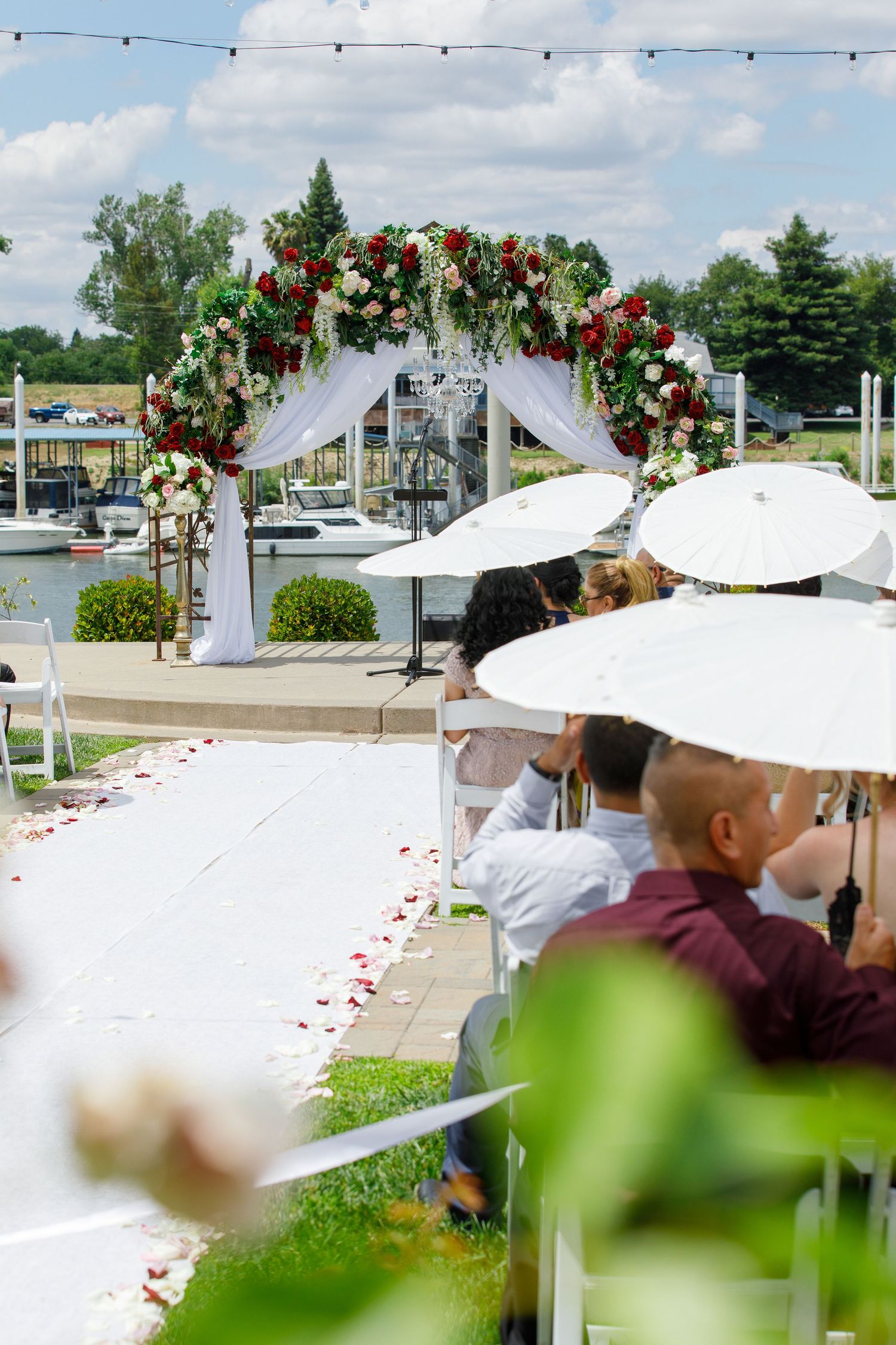 White parasols and floral arrangements line an outdoor wedding ceremony aisle with string lights overhead.