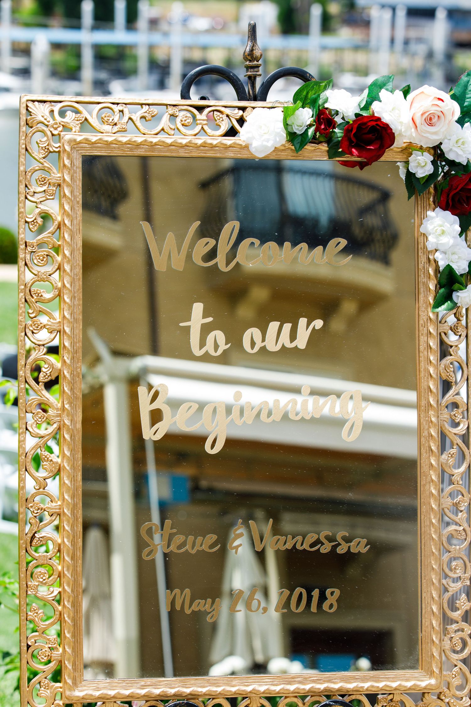 Elegant gold-framed mirror with 'Welcome to our Wedding' calligraphy text and floral decorations at an outdoor venue.