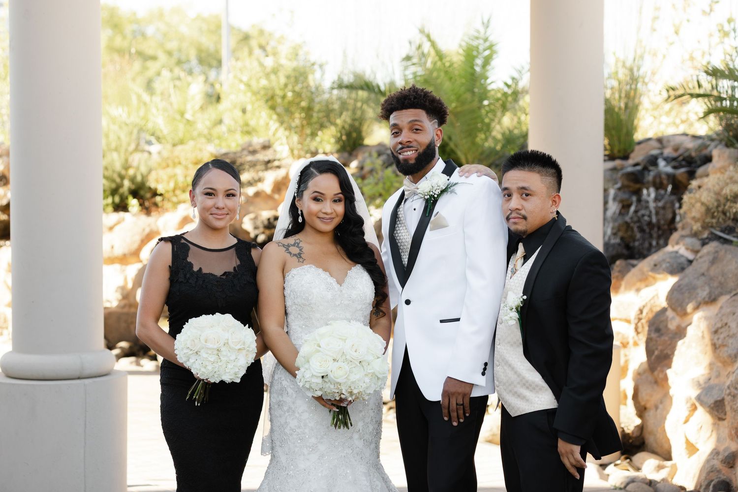 A wedding party poses together in formal attire under white columns during an elegant outdoor celebration.