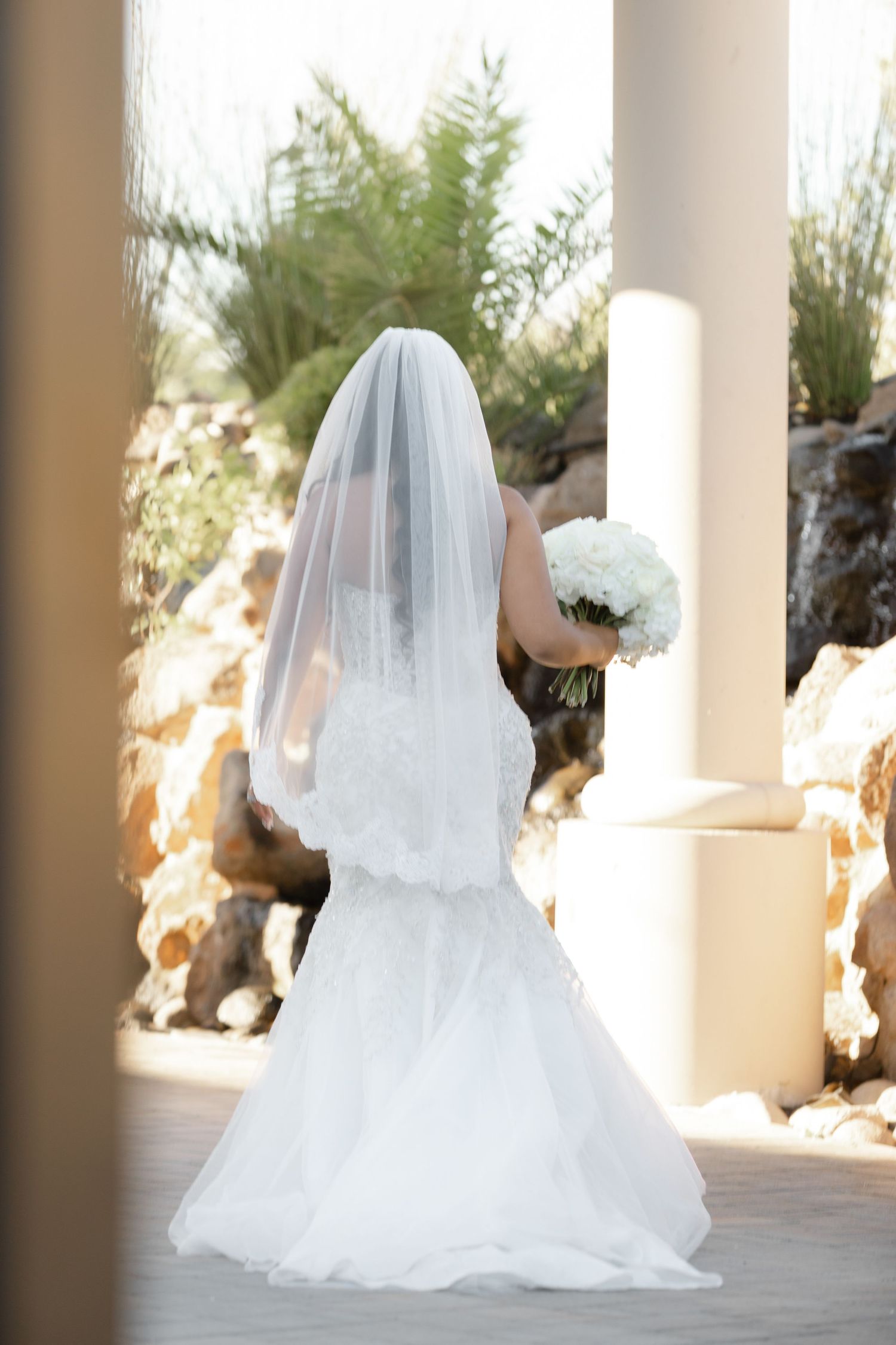Elegant bridal portrait showing flowing white wedding dress and veil against classic white columns in outdoor setting.