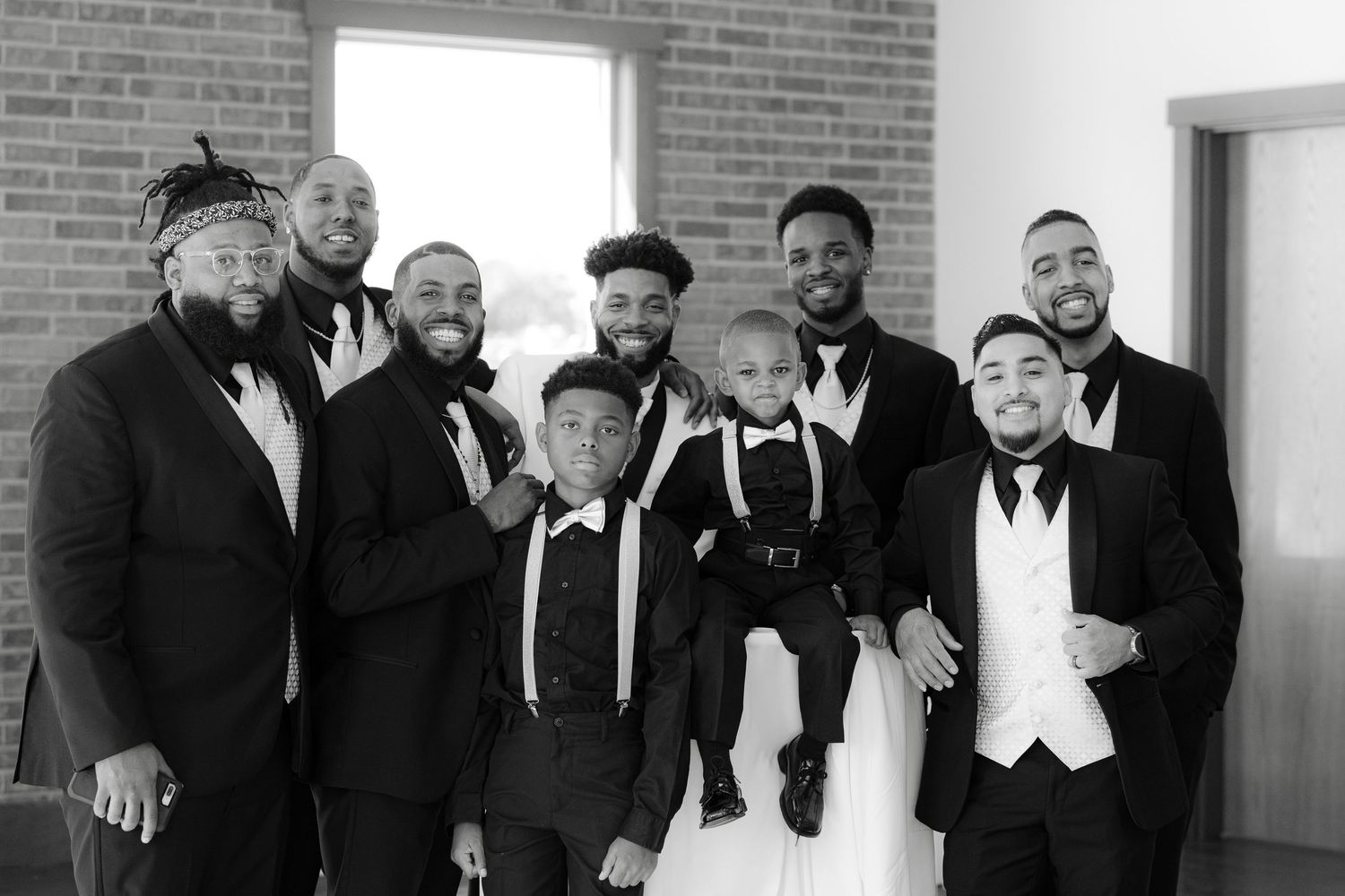 A group in formal black and white attire poses together against a brick wall in an interior setting.