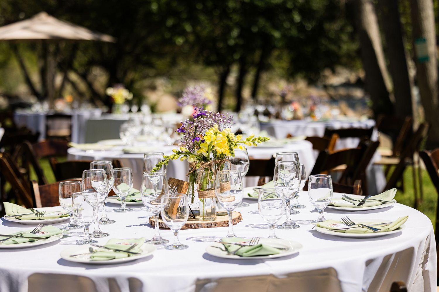 Elegant outdoor wedding reception table with white linens, floral centerpiece, and wine glasses set under trees.