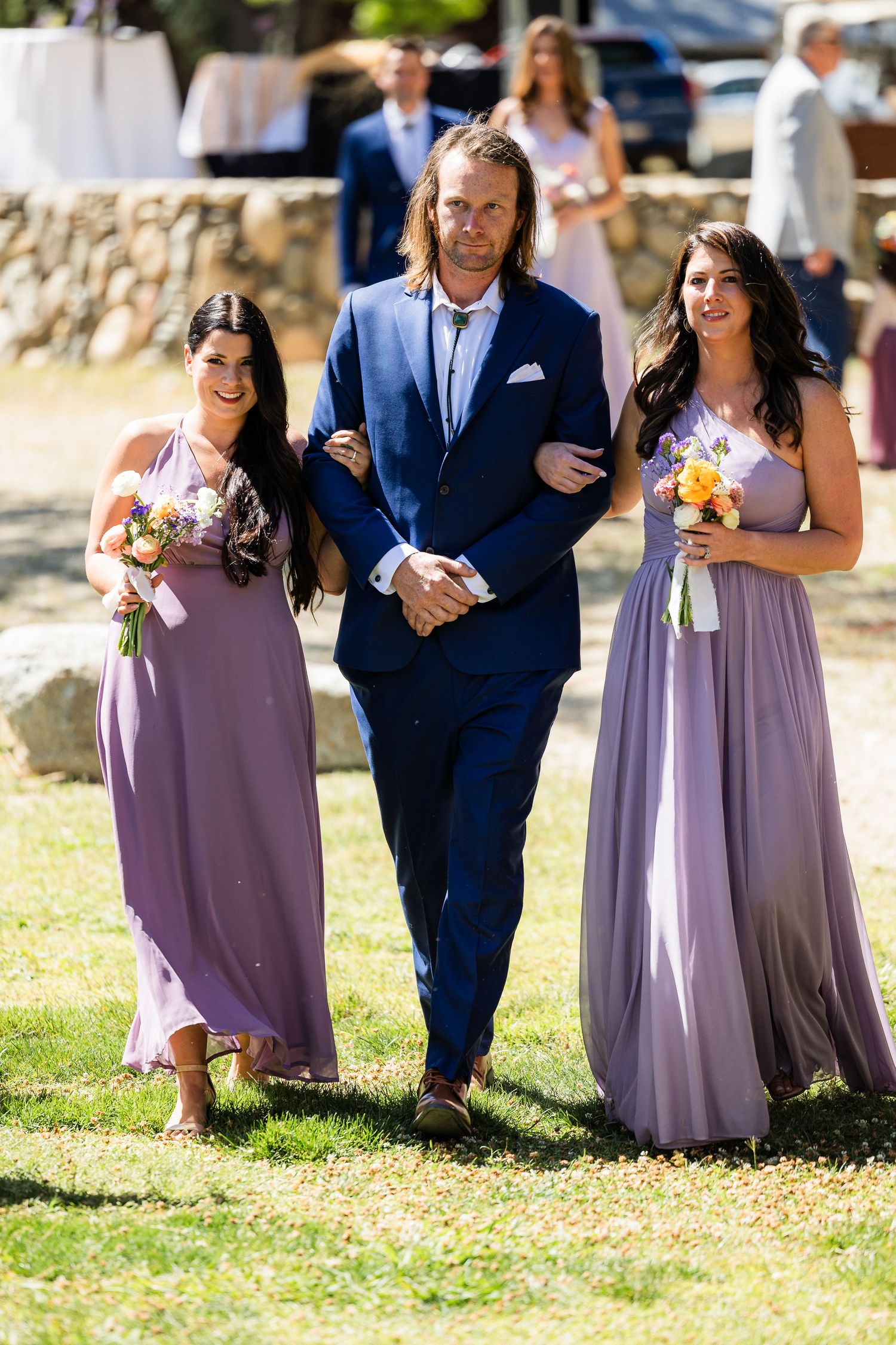 Wedding party members in navy suits and lavender dresses walk together on a sunny outdoor lawn.