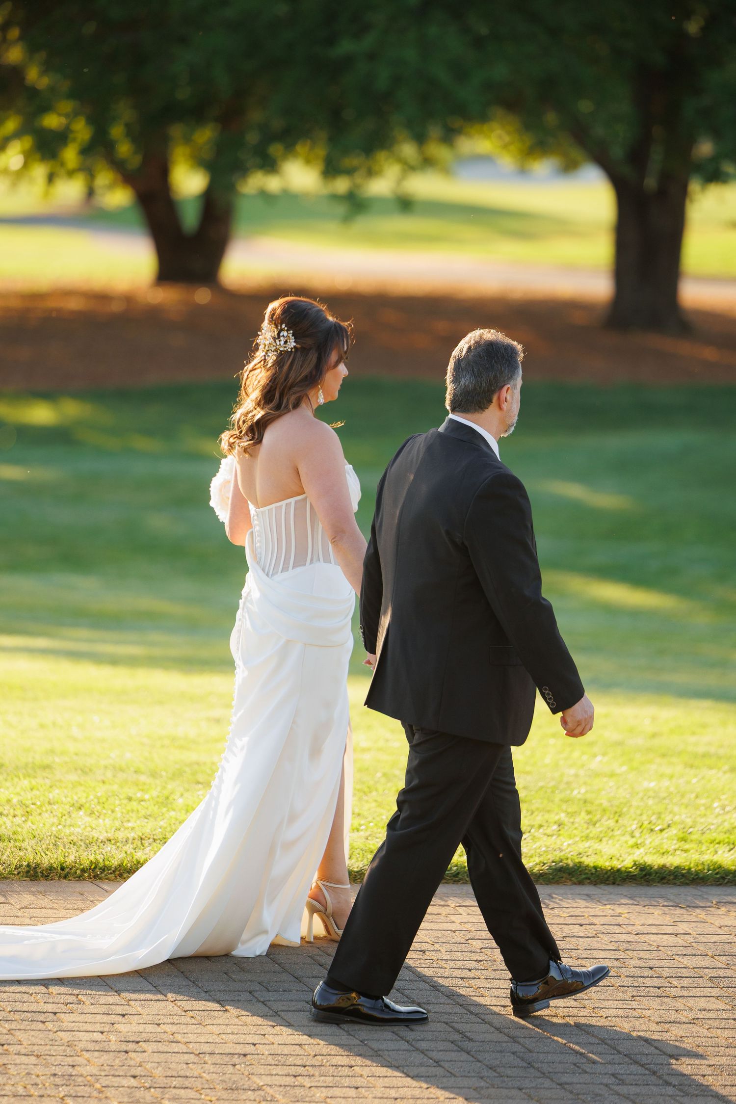 A bride in a flowing white gown and companion walk along a tree-lined path during golden hour.