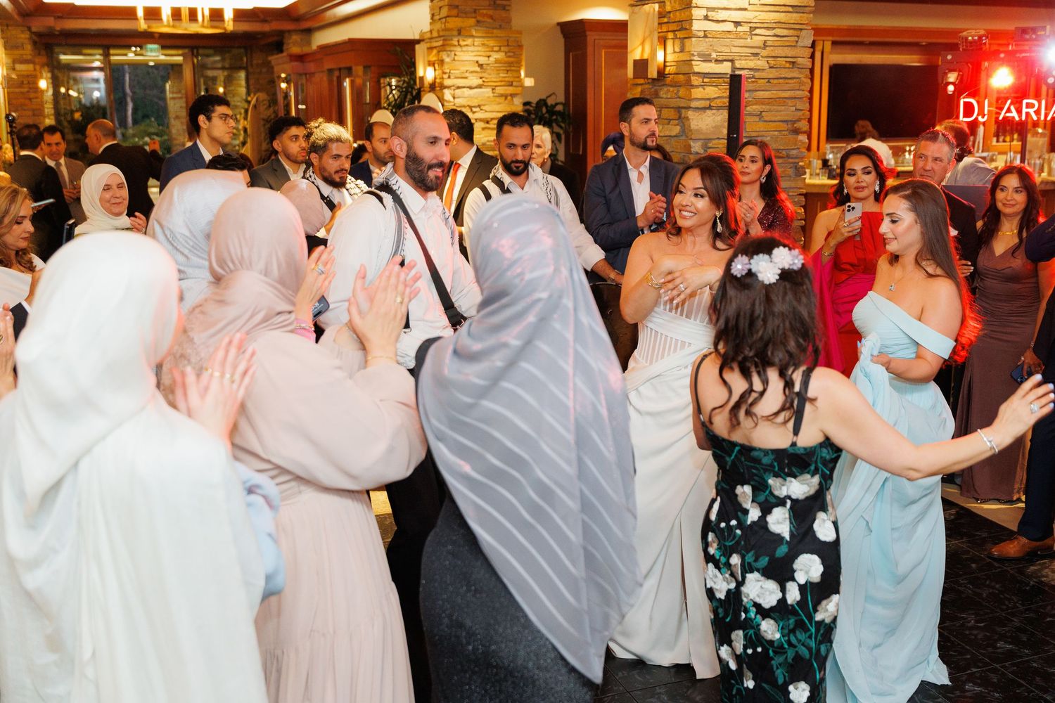 Wedding guests dance and celebrate together during a lively reception at a rustic venue with wooden walls and warm lighting.