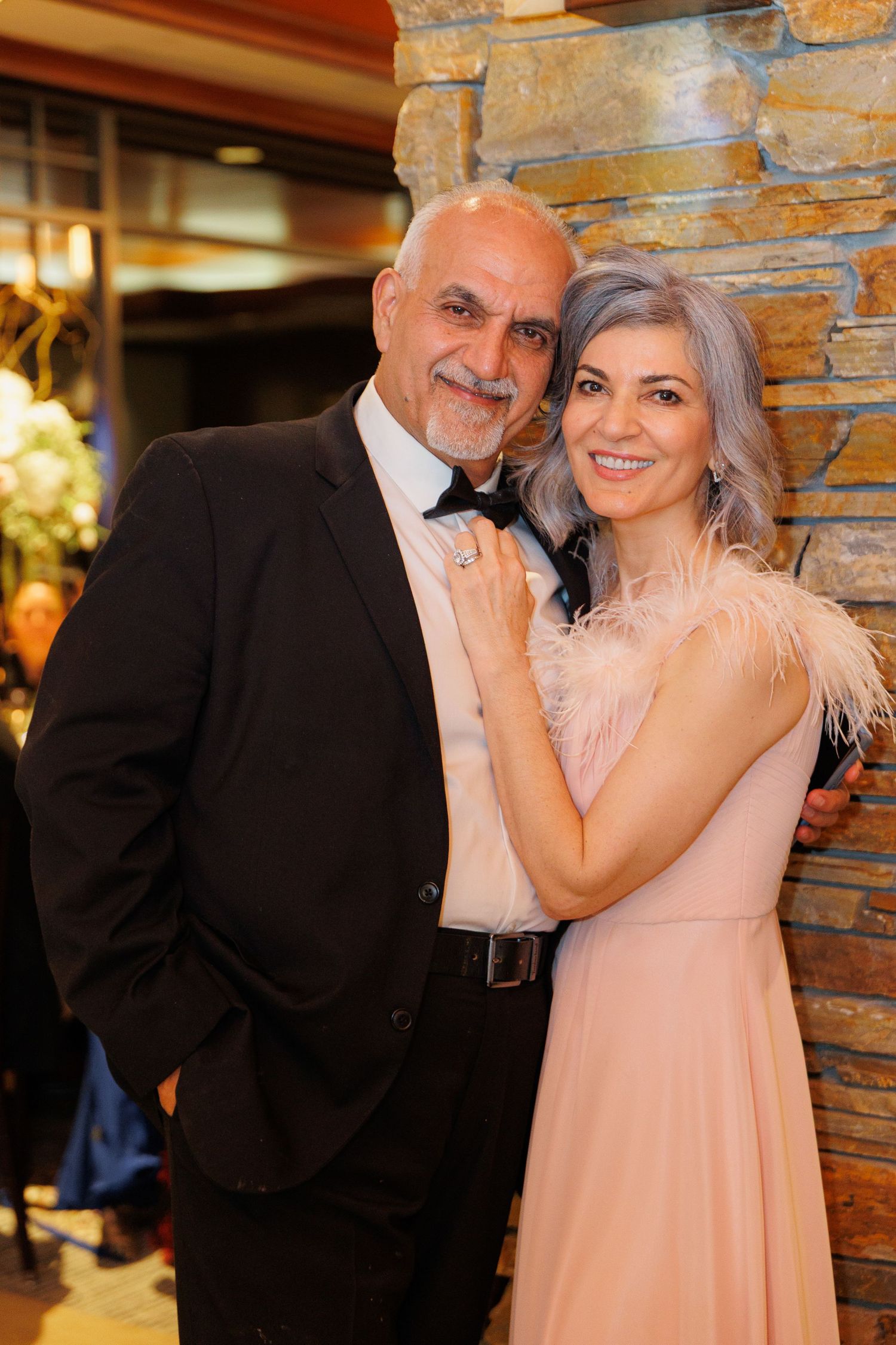 A couple in formal attire share tender moments against a rustic stone wall backdrop in a warm-lit venue.