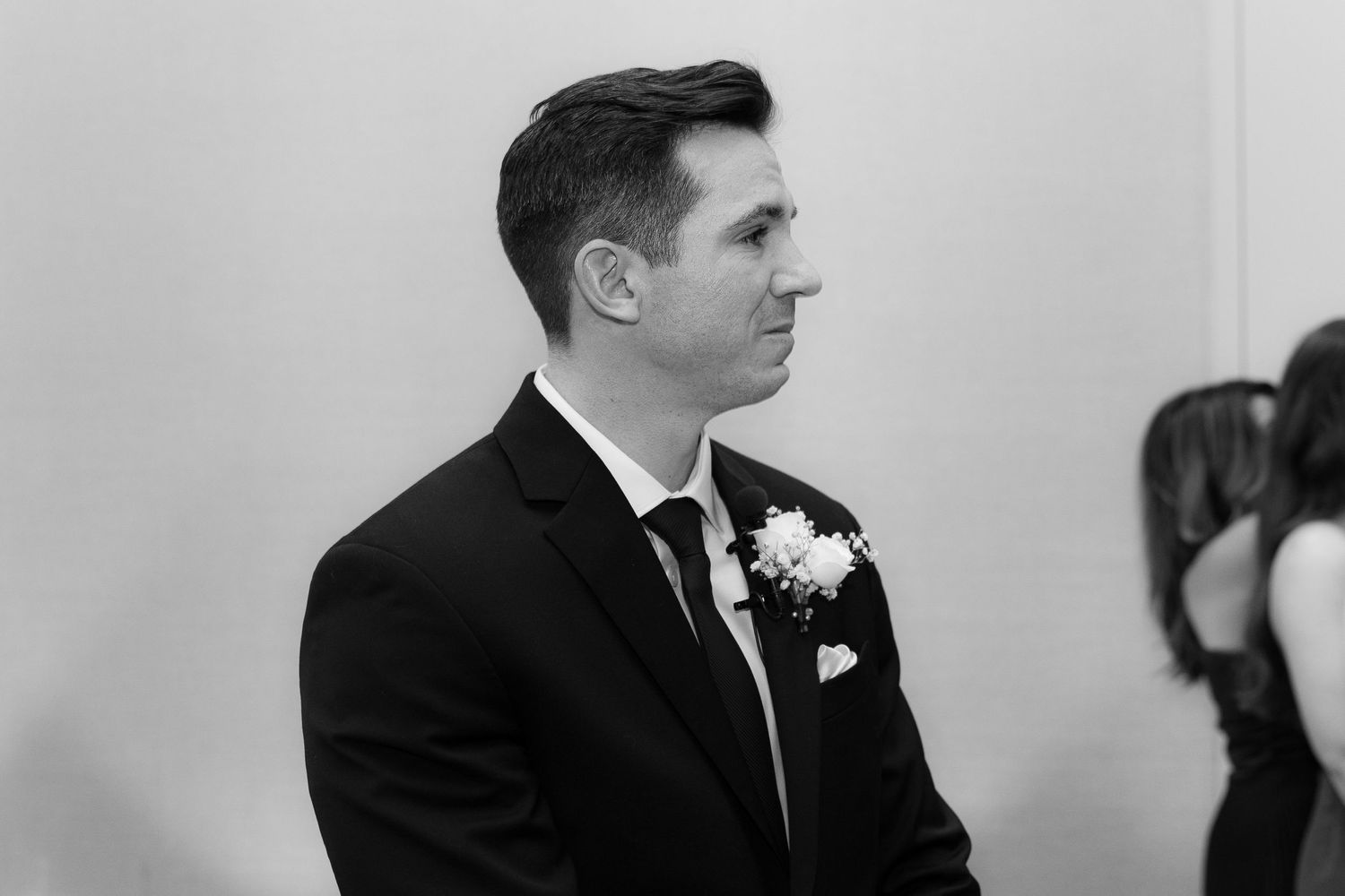 Black and white photo of a groom in a classic tuxedo with boutonniere looking to the side during wedding ceremony.