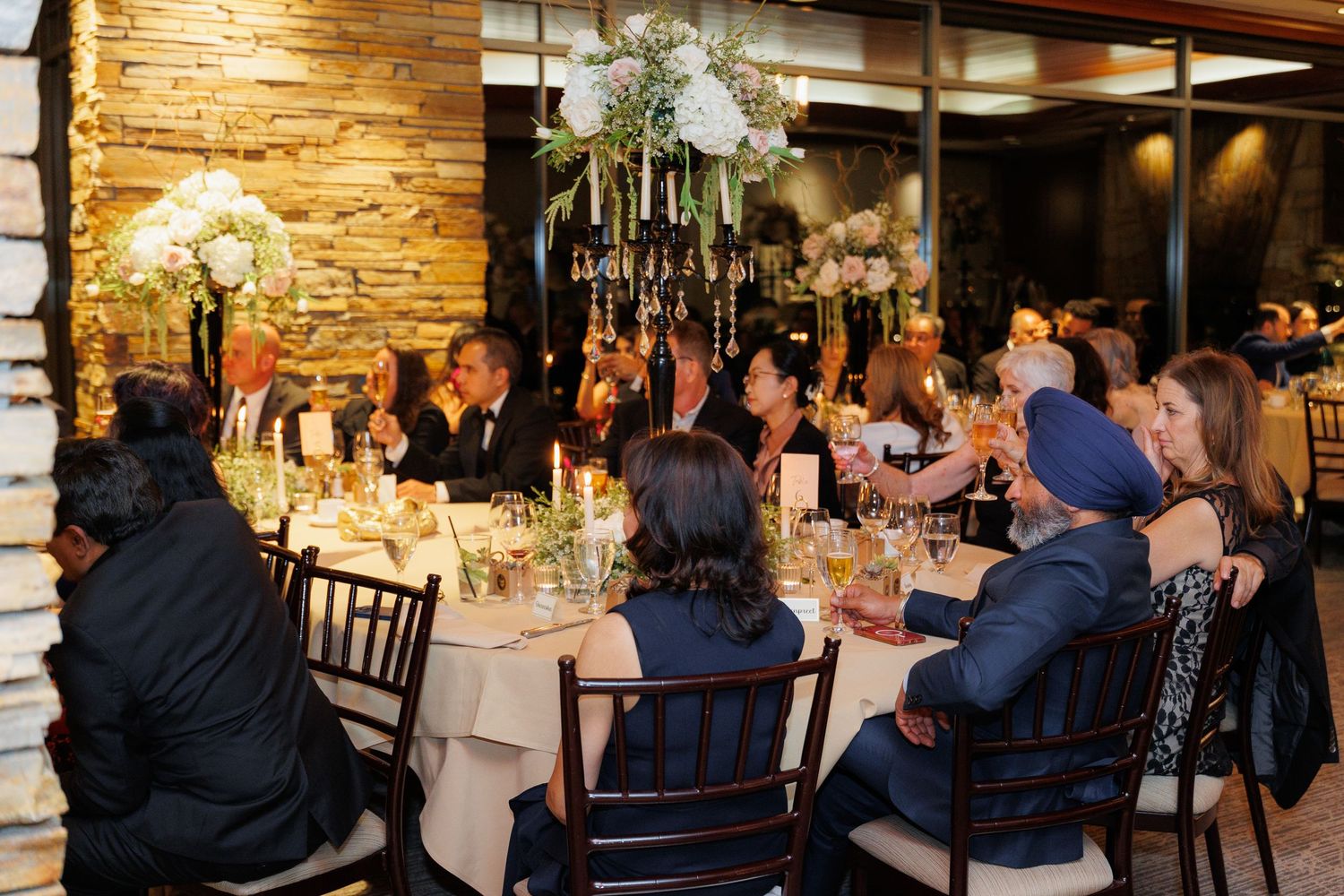 Elegant wedding reception dinner with white floral centerpieces and stone wall decor in a warmly lit restaurant setting.