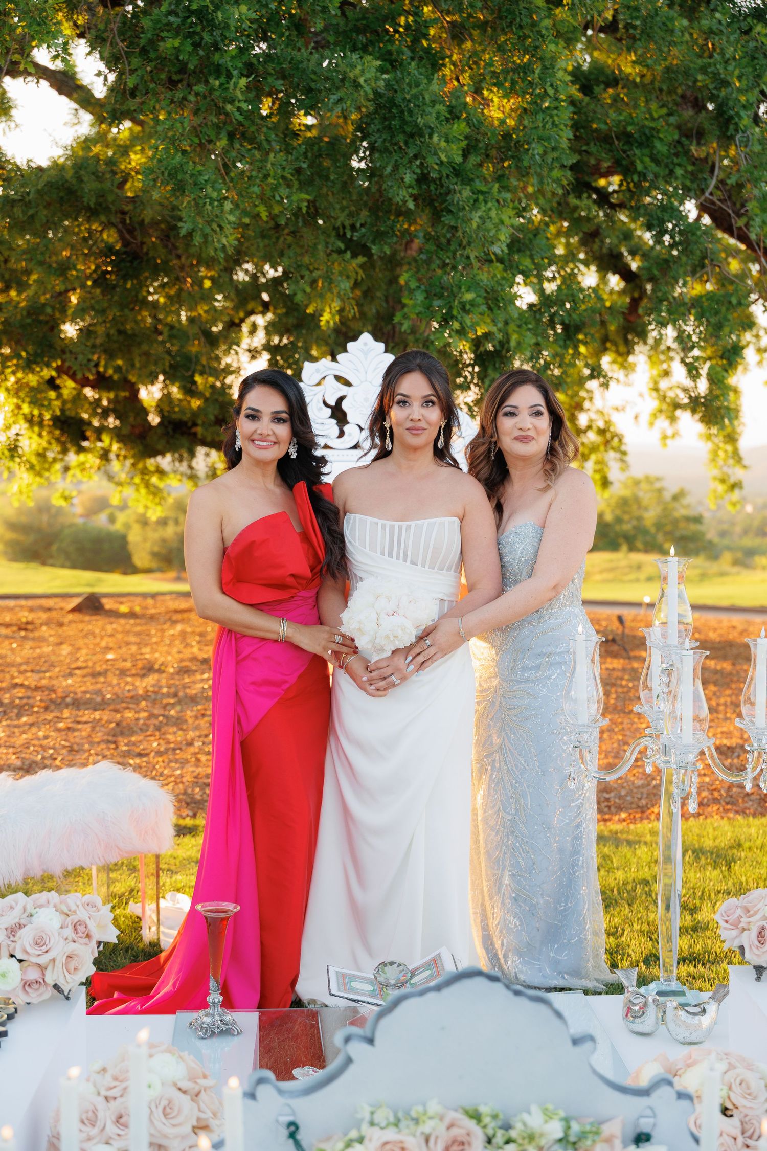 Wedding party in elegant dresses poses outdoors at sunset under trees with golden light filtering through branches.