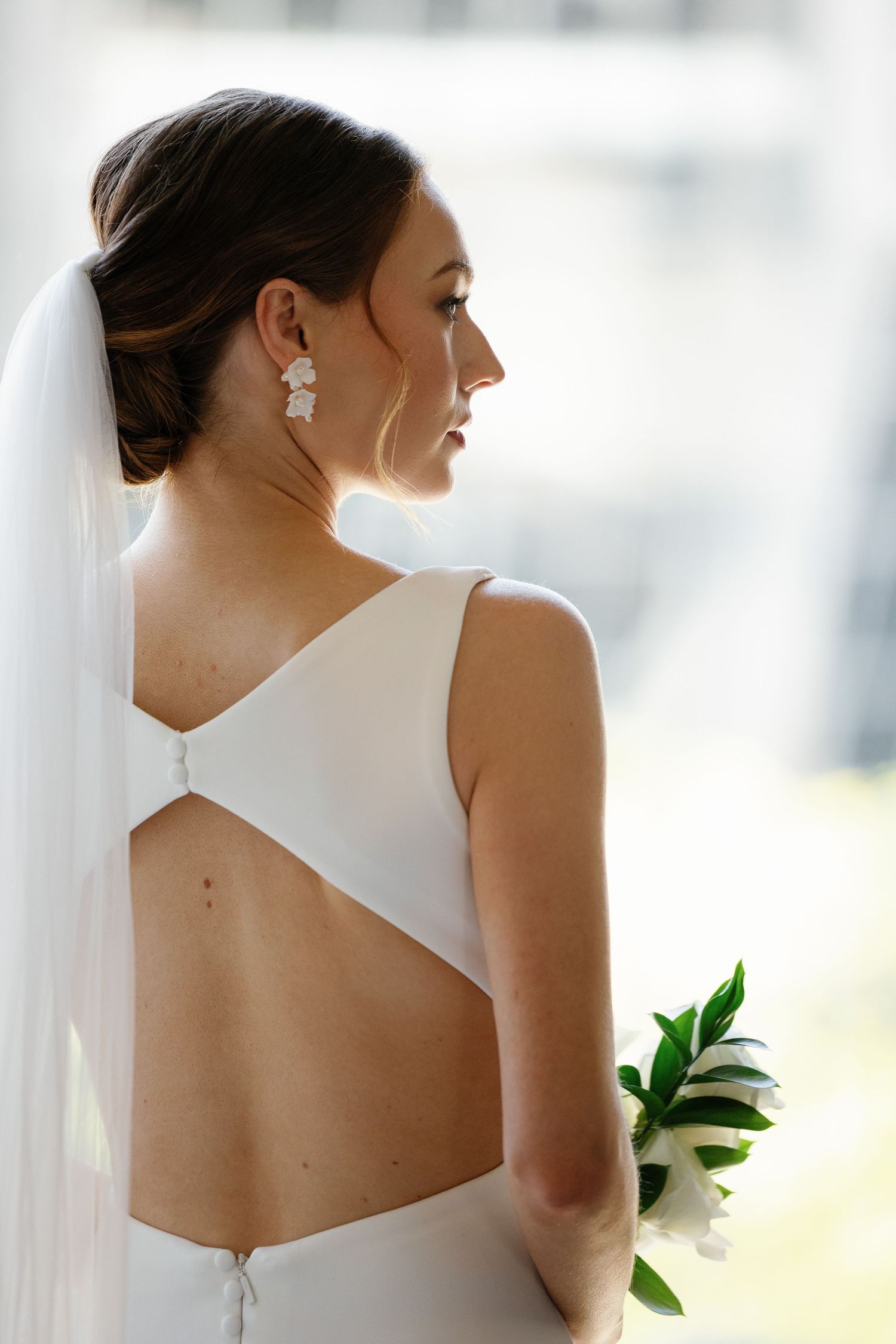Elegant bride in sleek white wedding dress with open back design and flowing veil against soft window light.