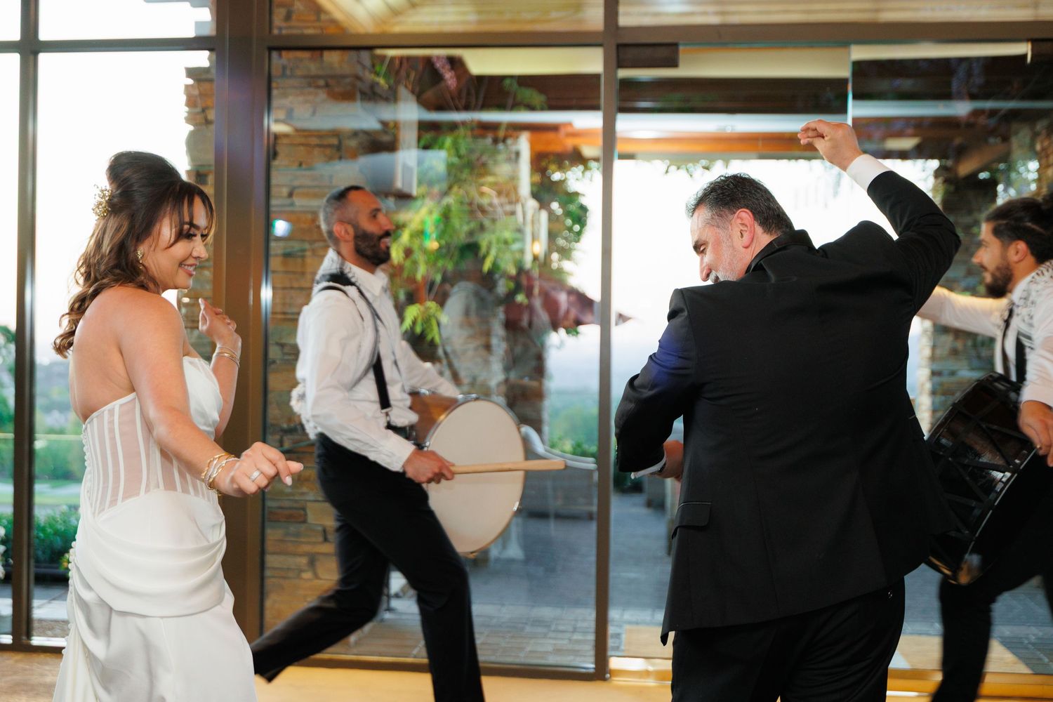 People dancing and celebrating at an indoor wedding reception with glass windows and natural lighting.
