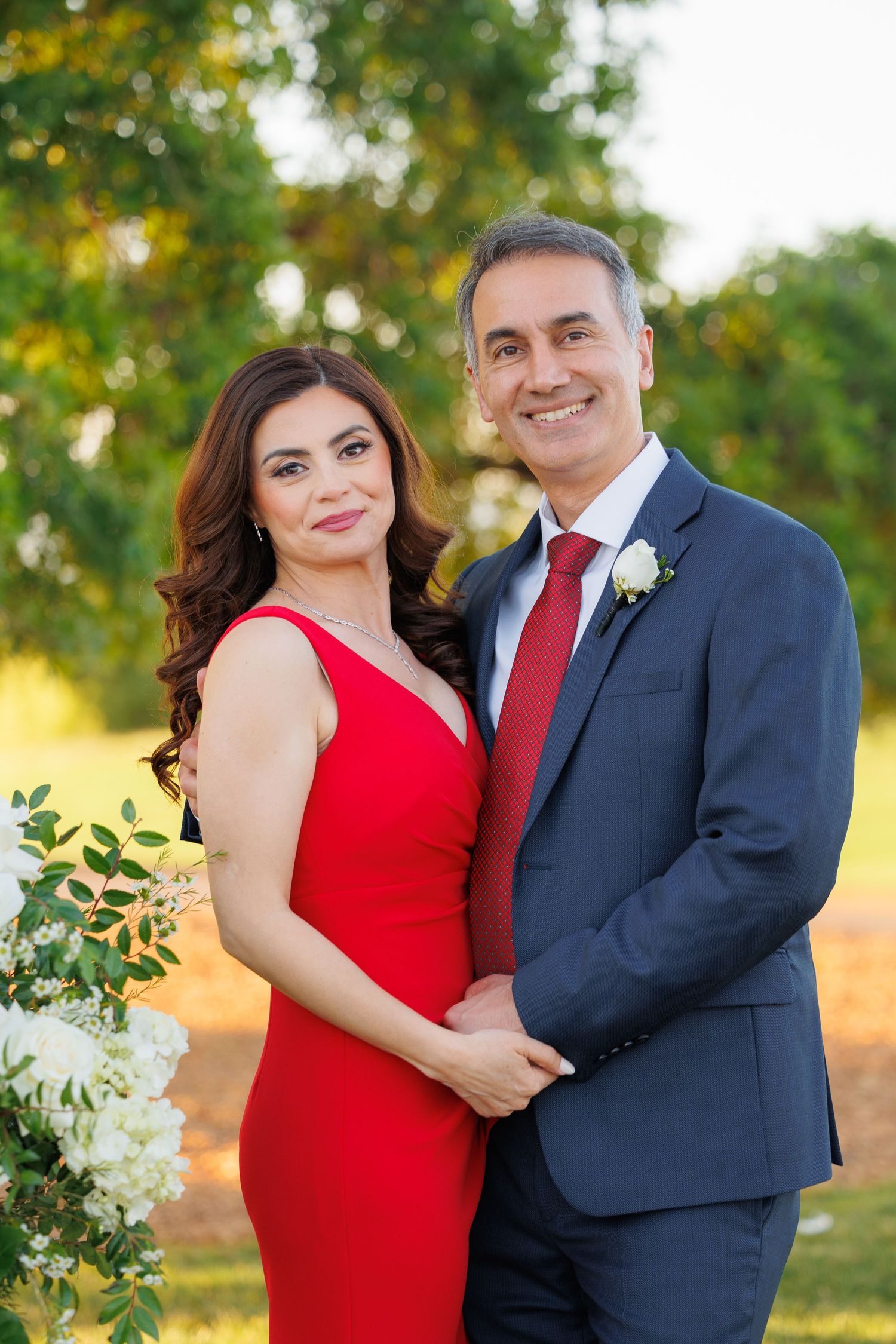 A couple poses outdoors in formal attire, with a red dress and navy suit against a sunlit natural background.