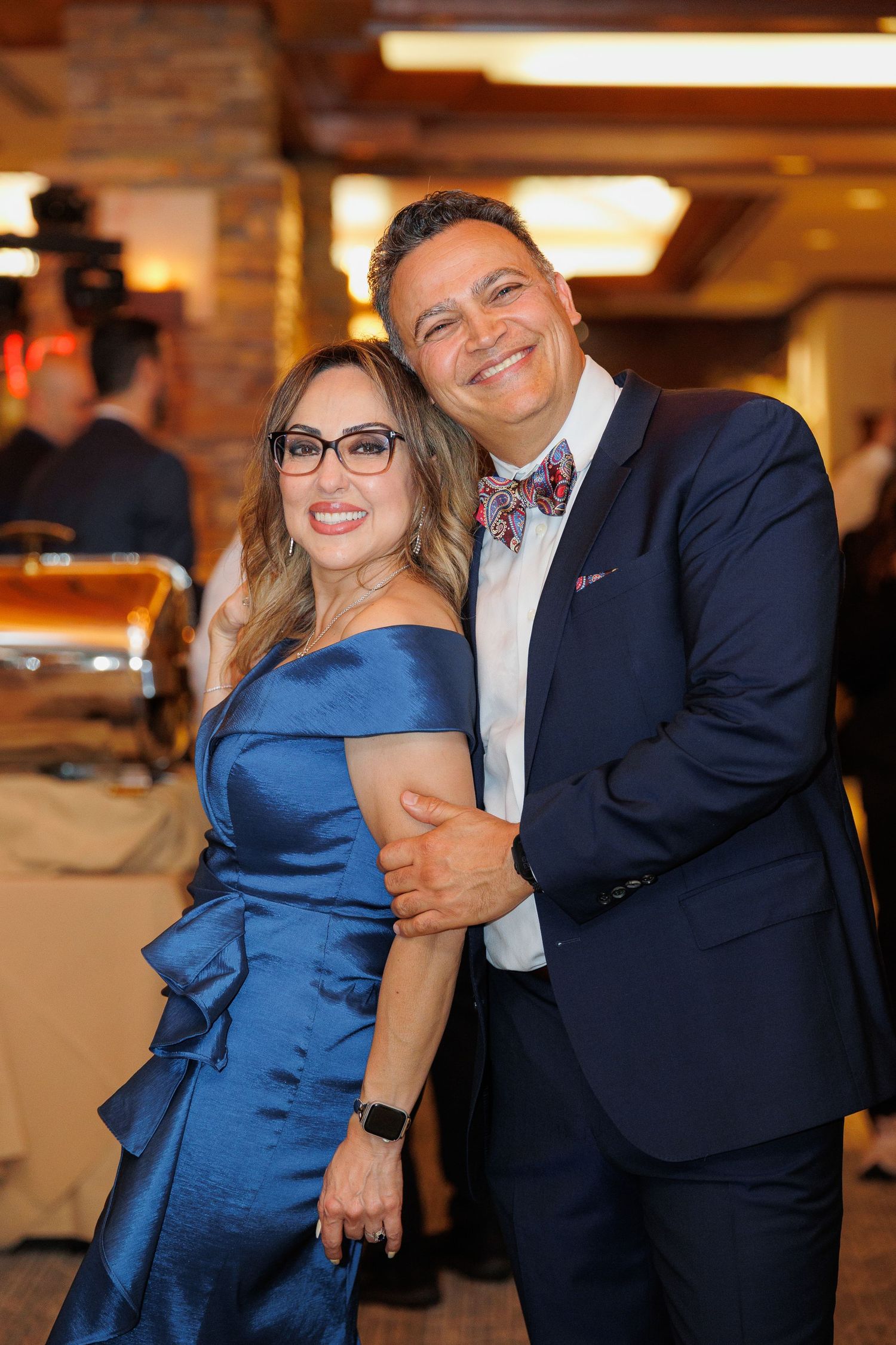 A couple in formal attire pose together at an event, with the woman in a blue satin dress and the man in a navy suit.