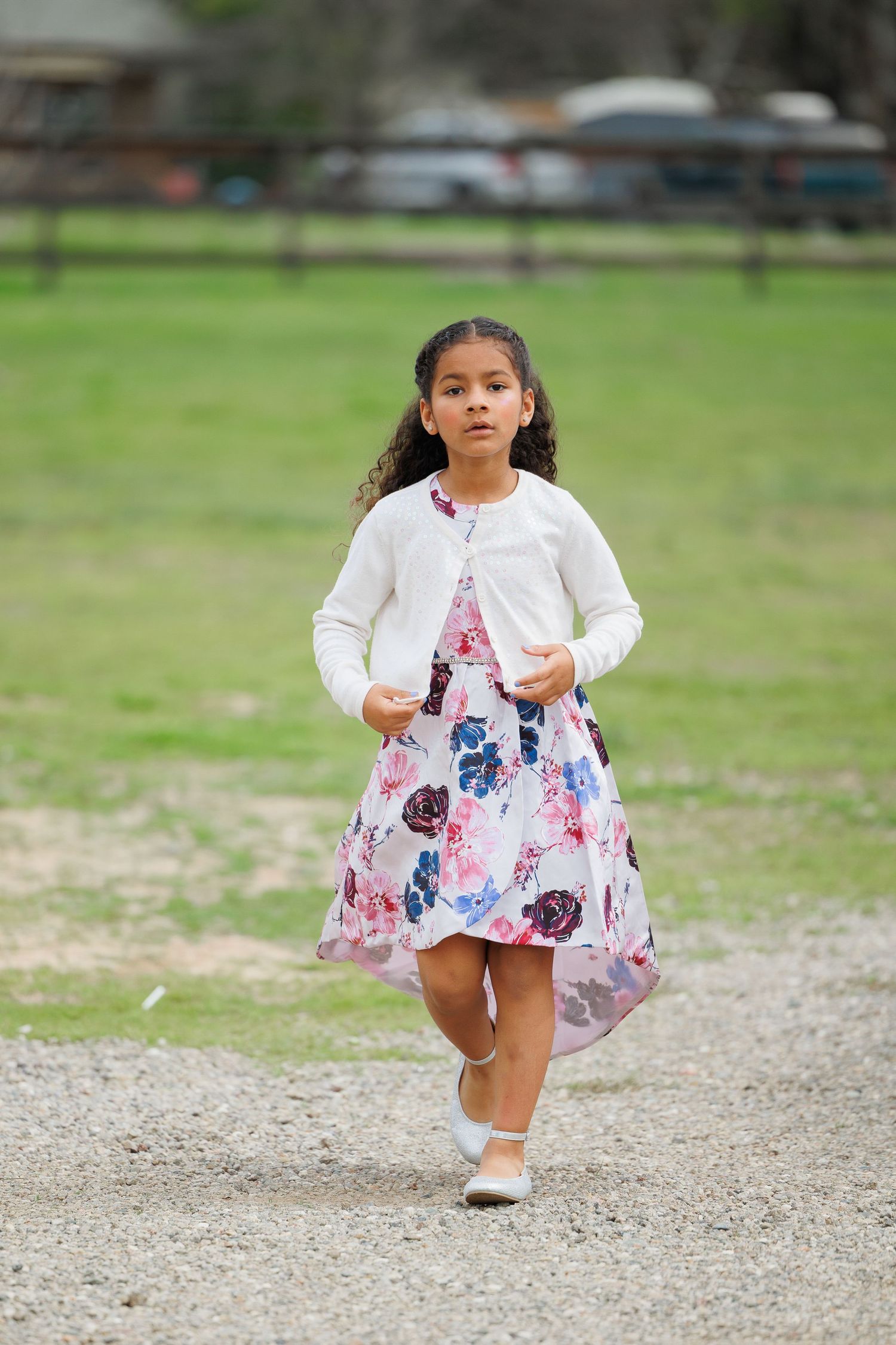 A young child in a white and floral dress skips playfully across a gravel path on a sunny day.