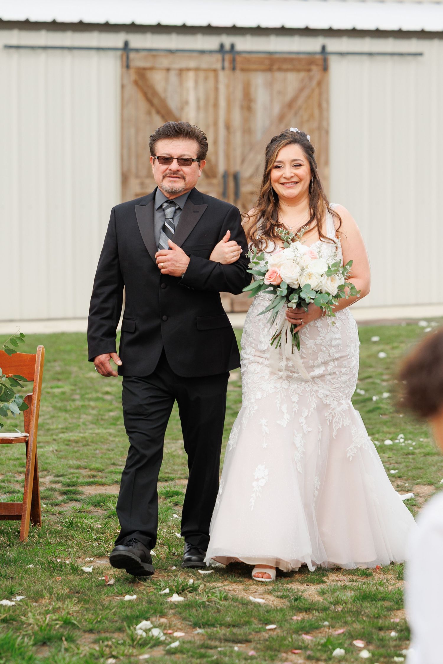 A bride in a lace wedding gown and companion in black suit walk arm-in-arm on grass in front of rustic barn door.