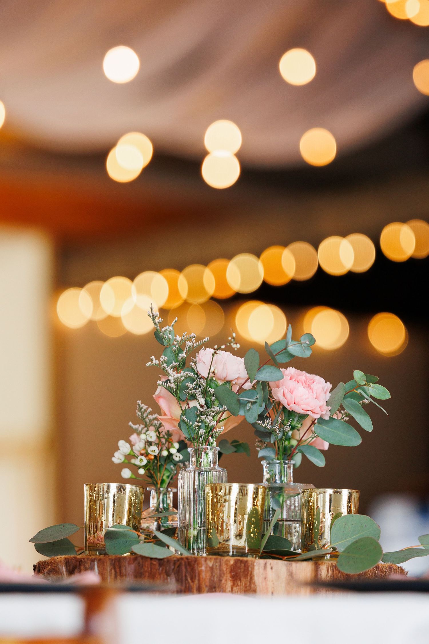 Small glass vases with pink roses and eucalyptus arranged on a wooden surface with twinkling bokeh lights in background.