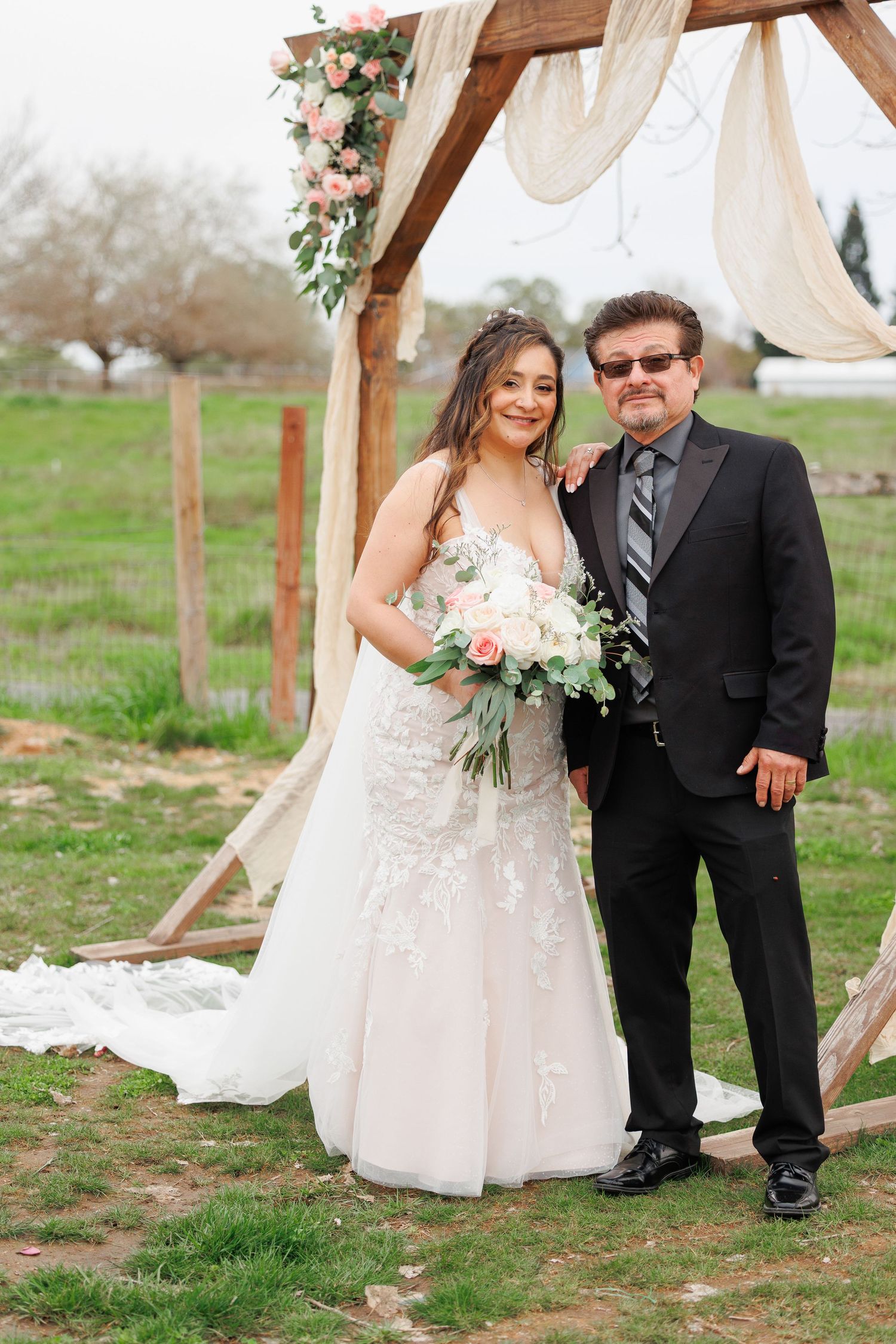 Couple poses together at rustic outdoor wedding ceremony with floral arch and draped fabric decor.