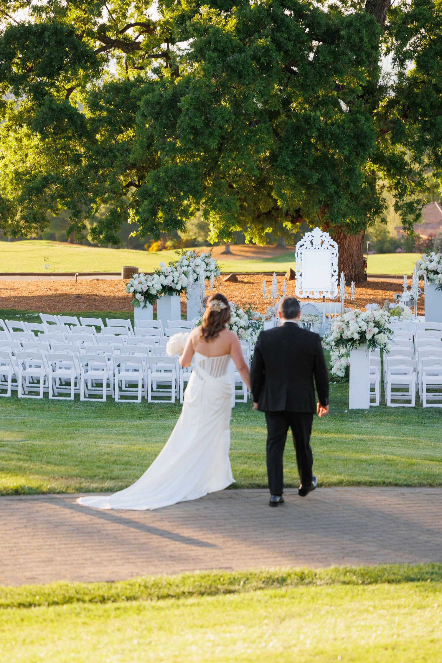 A couple walks along a paved path at an outdoor wedding venue with white chairs set up on the lawn at sunset.