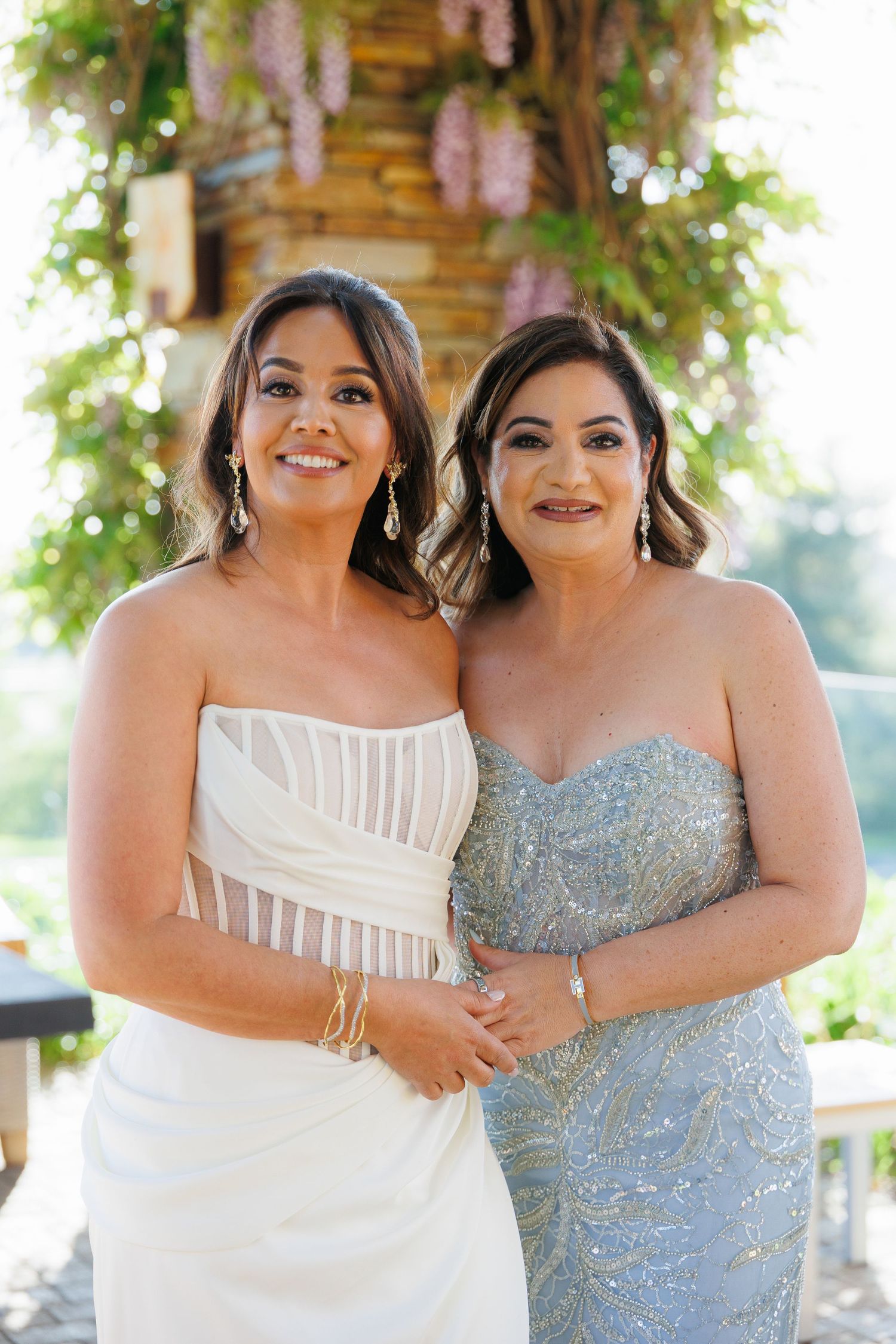 Two women in elegant strapless gowns pose together under floral decorations at a formal event.