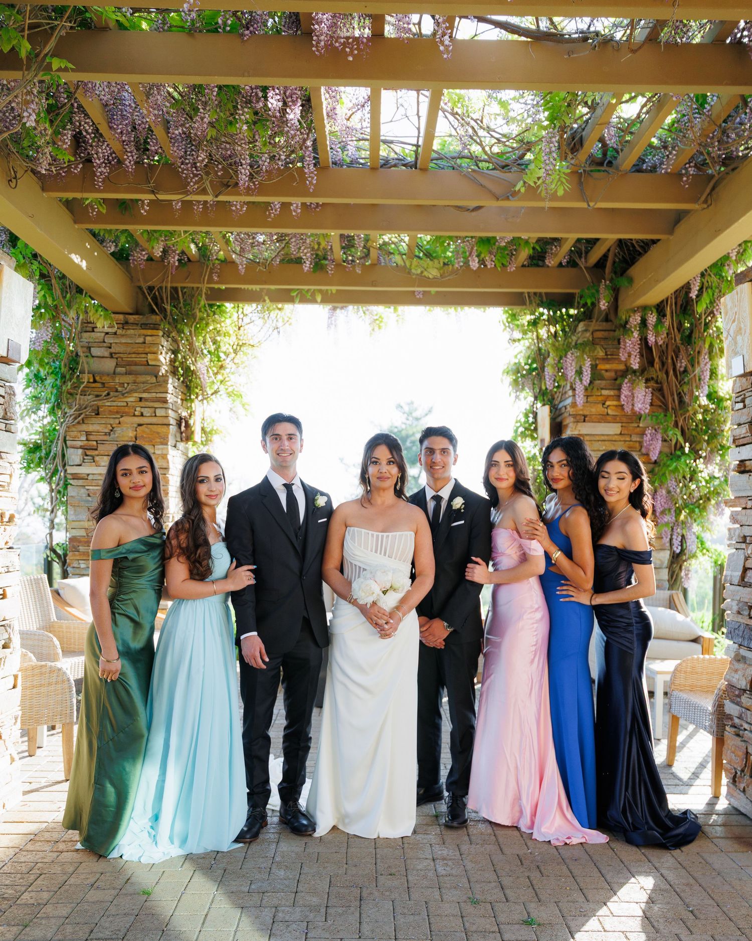 Wedding party poses together under a rustic pergola decorated with lush greenery and flowers in an outdoor venue.