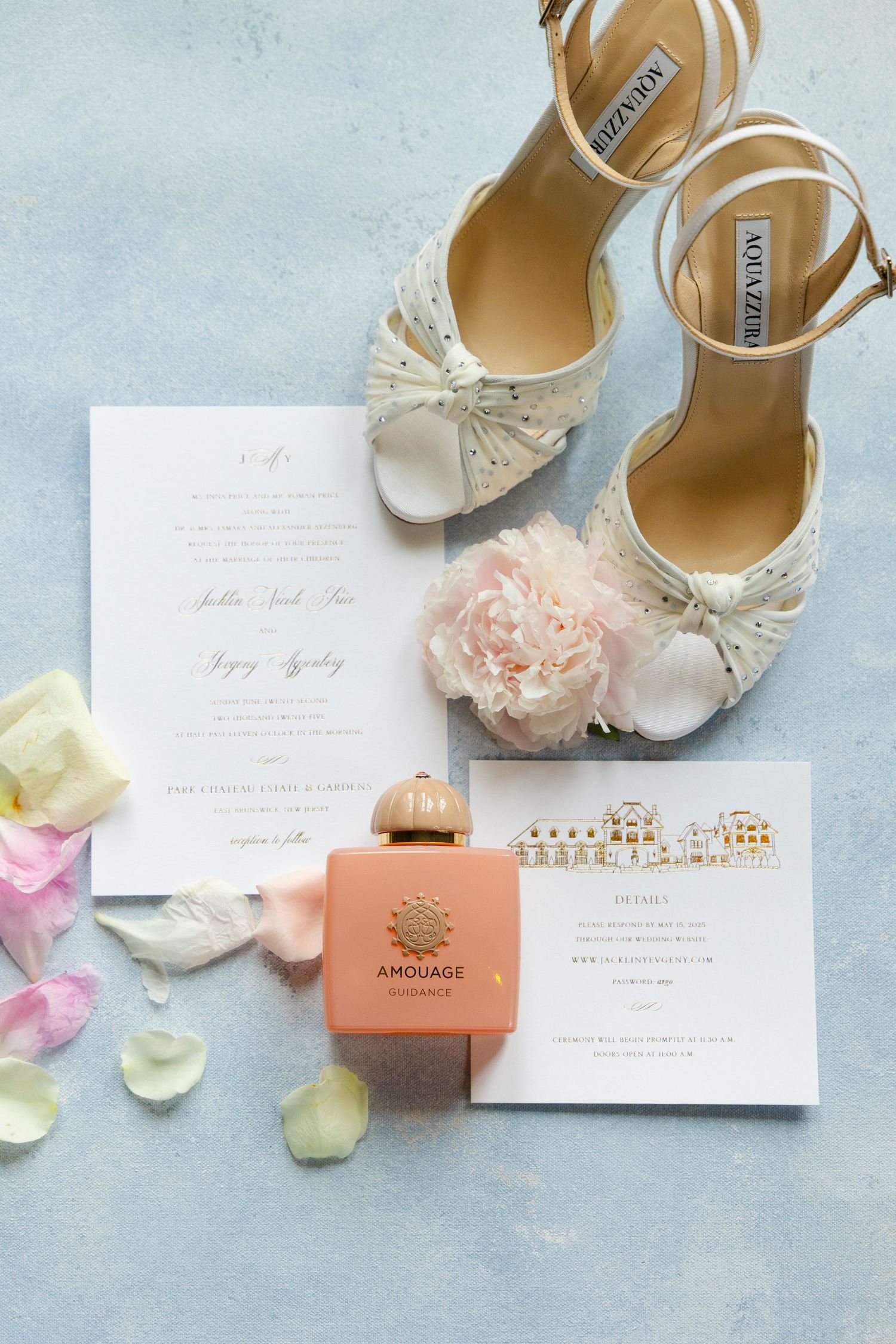 Wedding details flatlay with elegant bridal shoes, invitation cards, perfume bottle and scattered rose petals on blue surface.